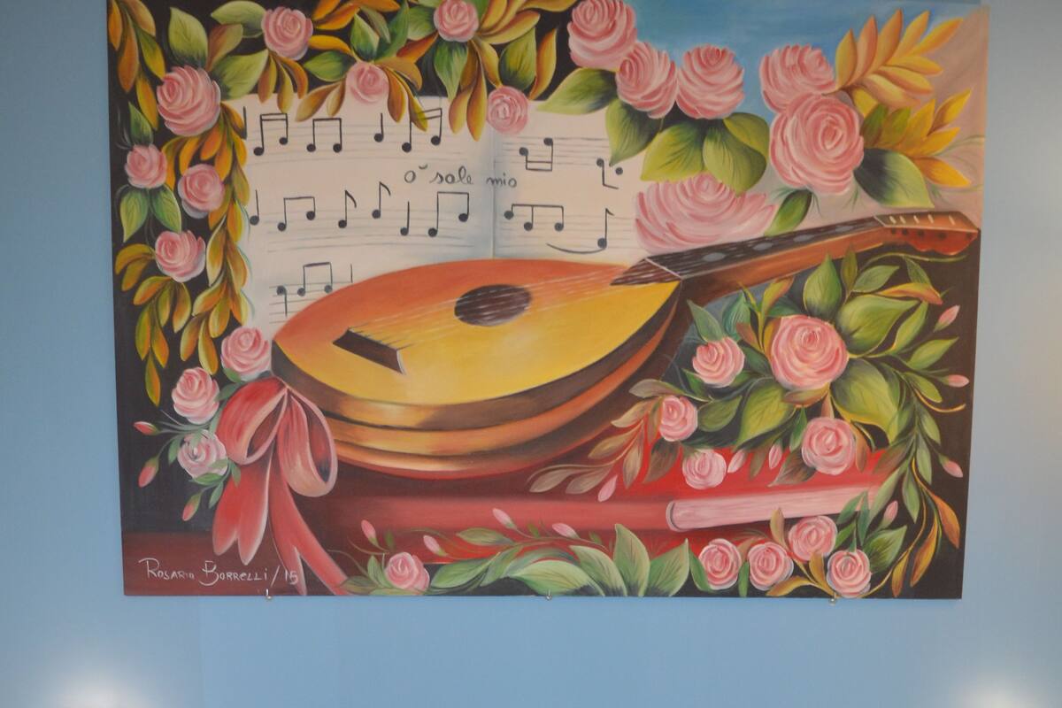 A hotel room with a bed, a painting of musical instruments and flowers, and a bedside table with a lamp.