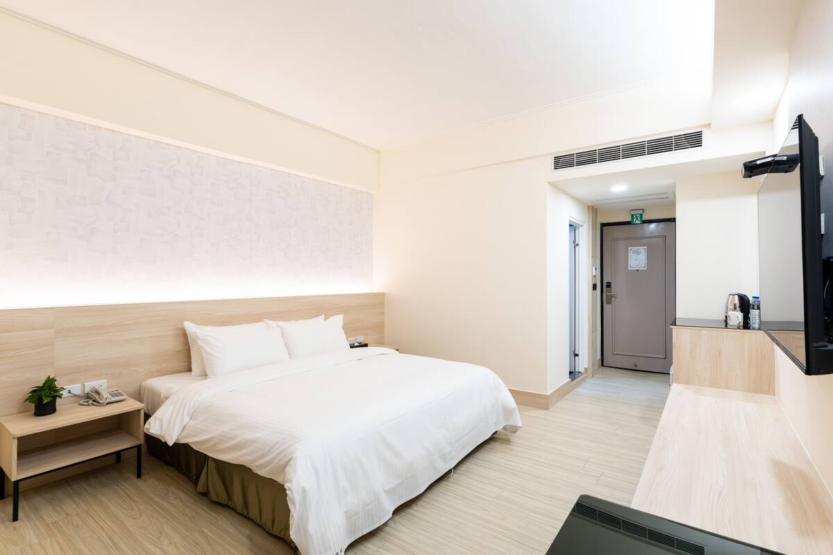 A hotel room with a bed, bedside table, and a small plant. There is a door leading to a bathroom and a kitchenette area.