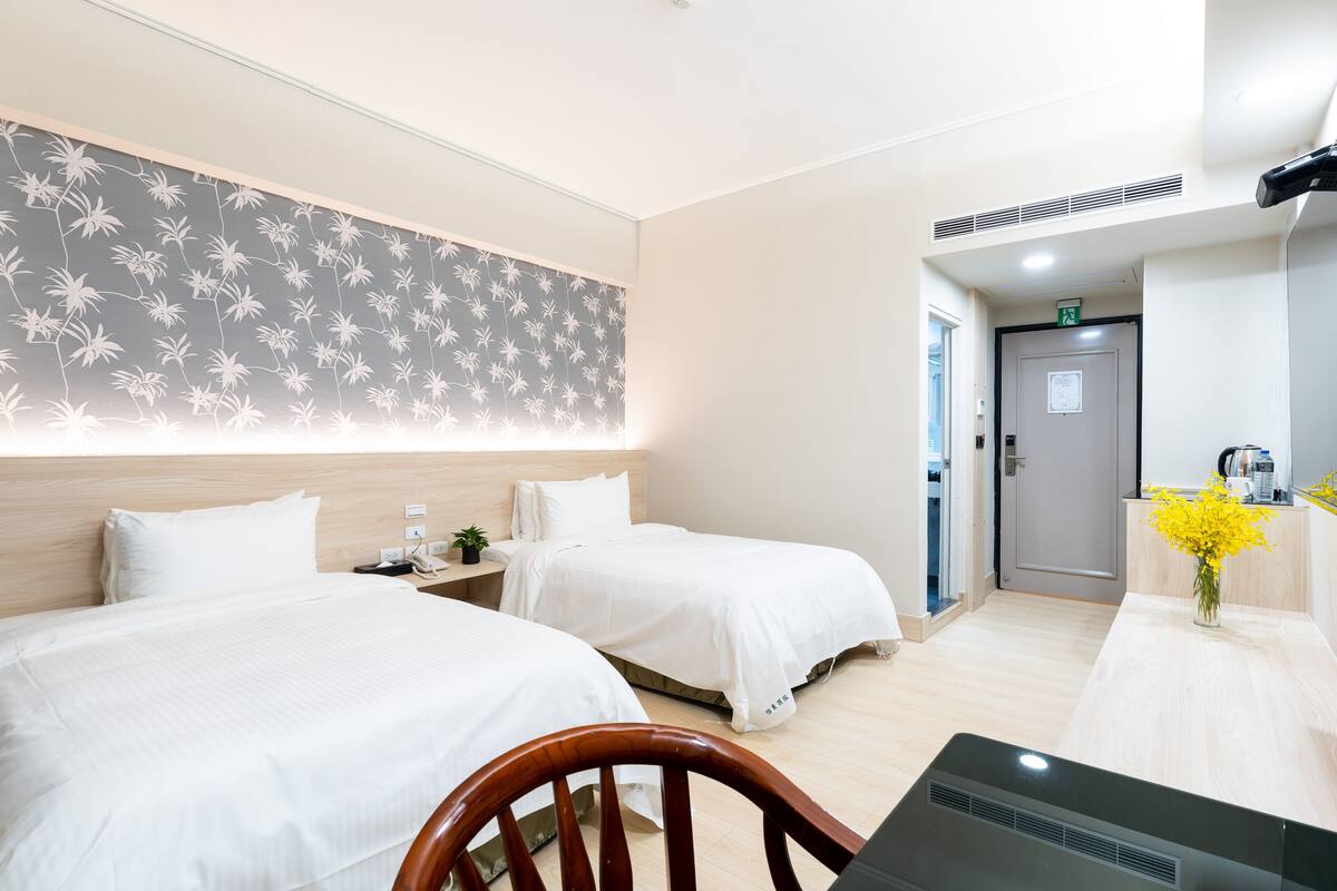 A hotel room with two beds, a desk, a chair, and a small table with a vase of flowers.