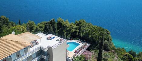 A high-rise building with a rooftop pool, surrounded by trees and overlooking a clear blue sea.