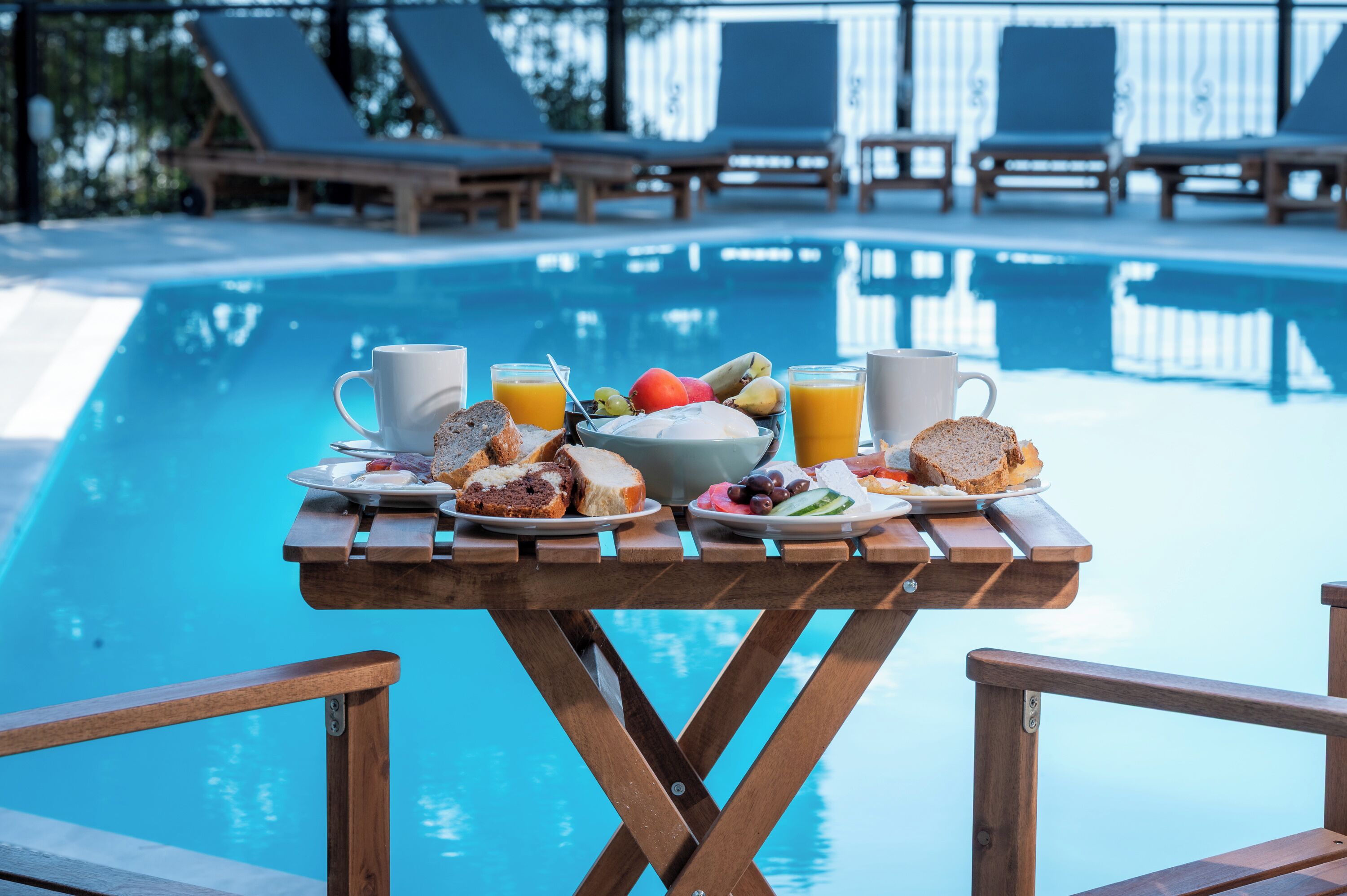 Mesa de desayuno junto a la piscina con platos de comida, bebidas y vistas a la piscina y tumbonas.