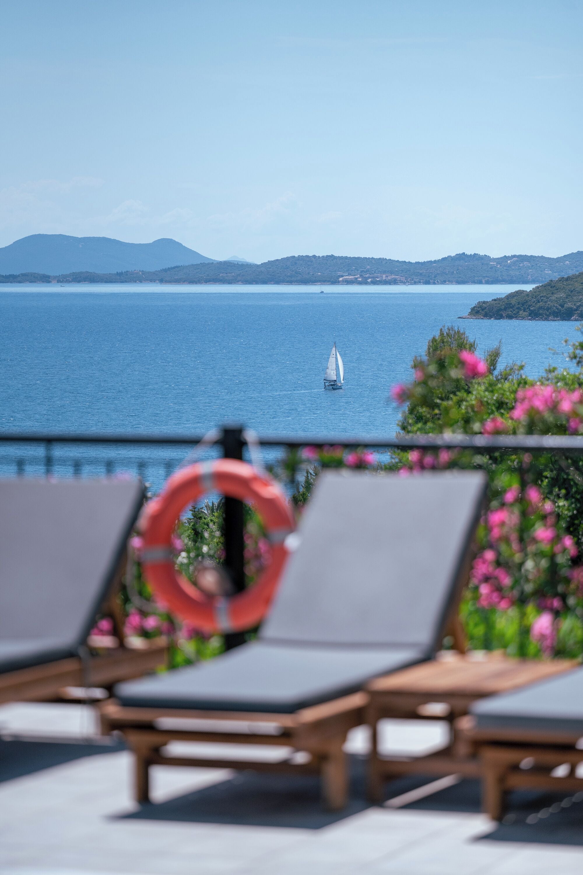 A seaside view with lounge chairs, a life ring, and a sailboat on the water.