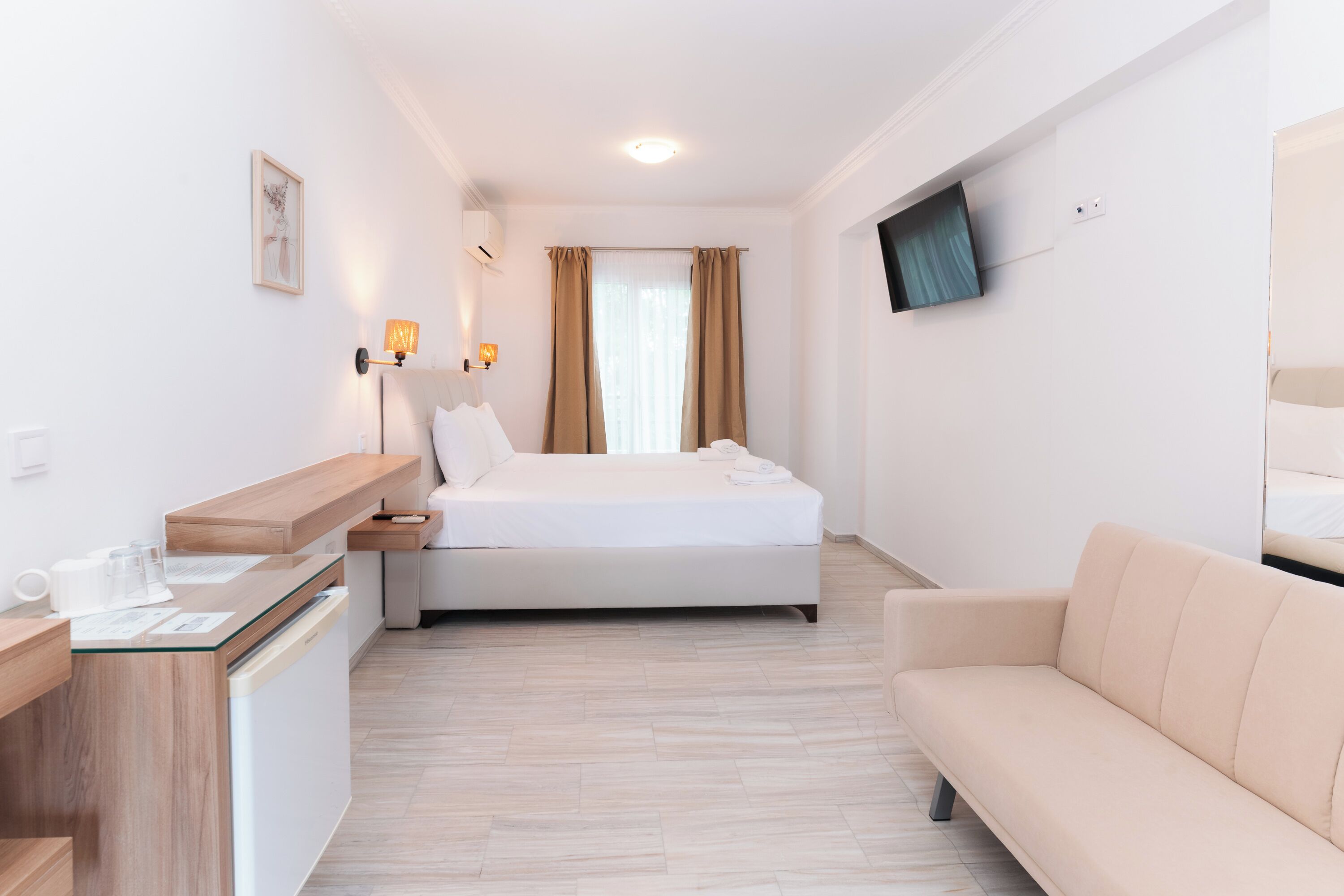 A hotel room with a bed, a sofa, a desk with a glass top, a TV mounted on the wall, and a window with curtains.