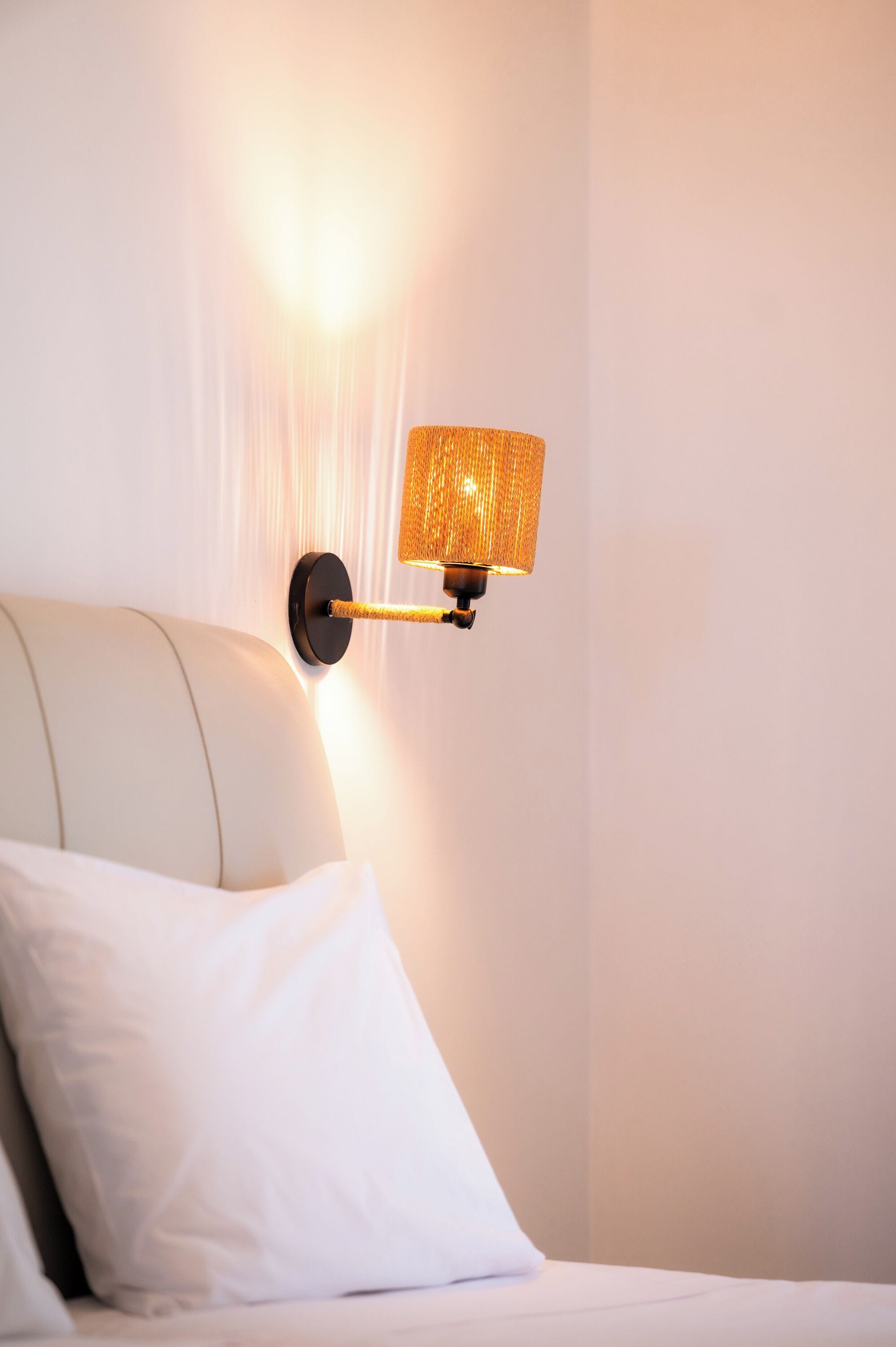 A wall-mounted lamp with a textured shade, a white pillow, and a bed with a headboard.