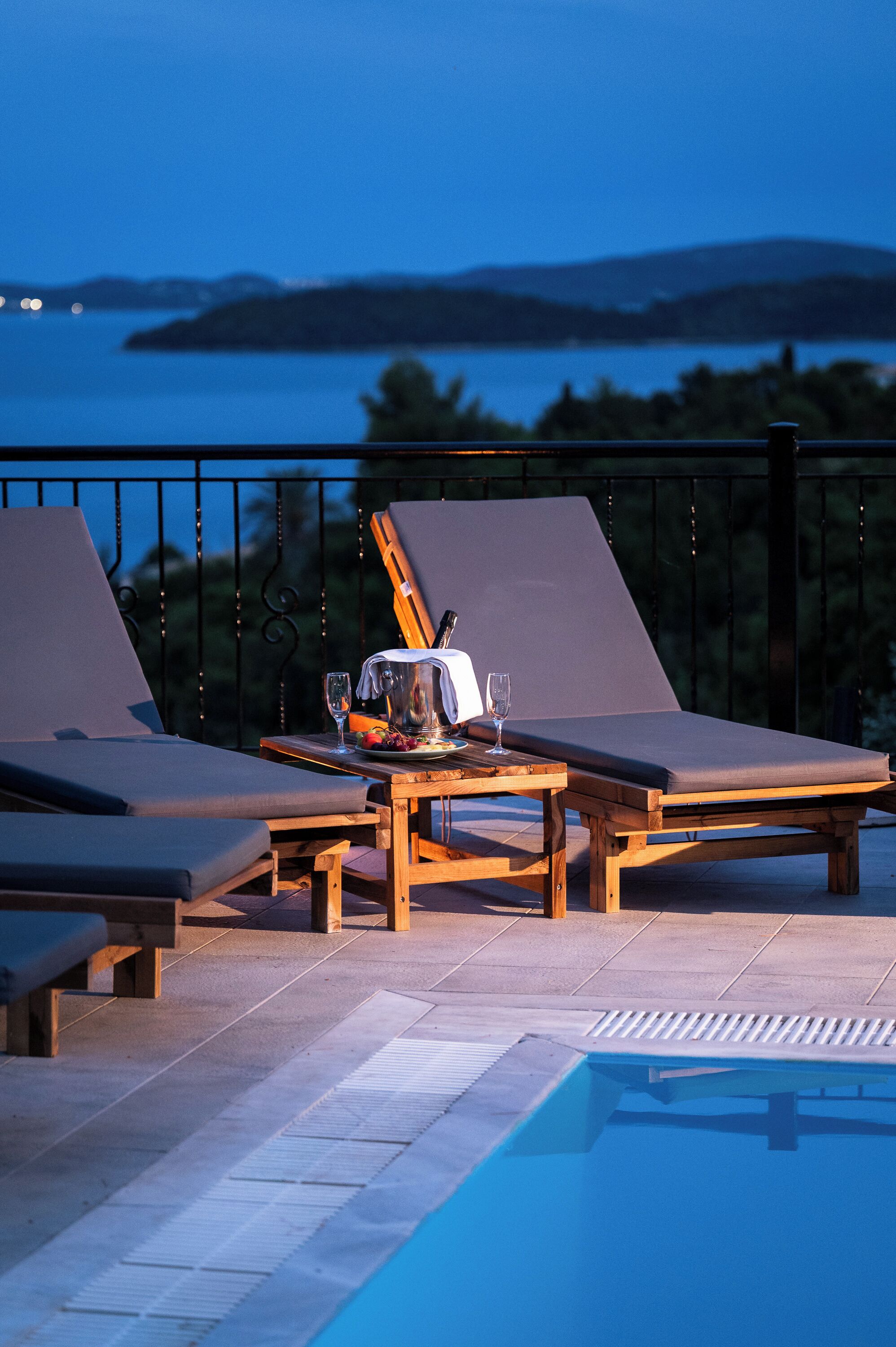 Uma área à beira da piscina com espreguiçadeiras, uma mesa com jarra e copos, e vista para a água e terra ao longe.