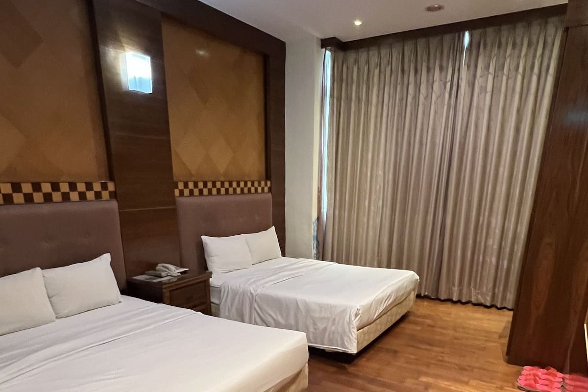 A hotel room with two beds, wooden flooring, a large window with curtains, and a small table with a phone.