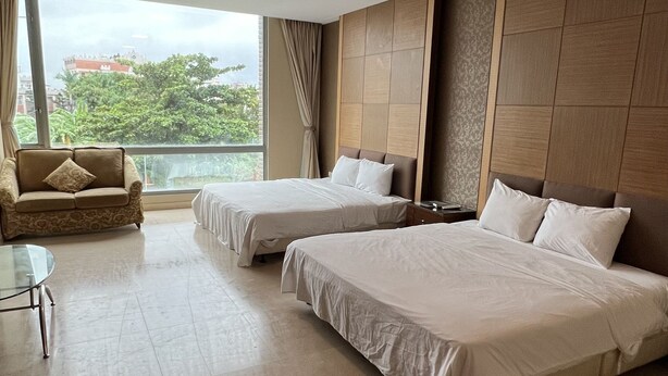 Habitación de hotel con dos camas, un sofá, una mesita y ventanas amplias con vistas a la ciudad.