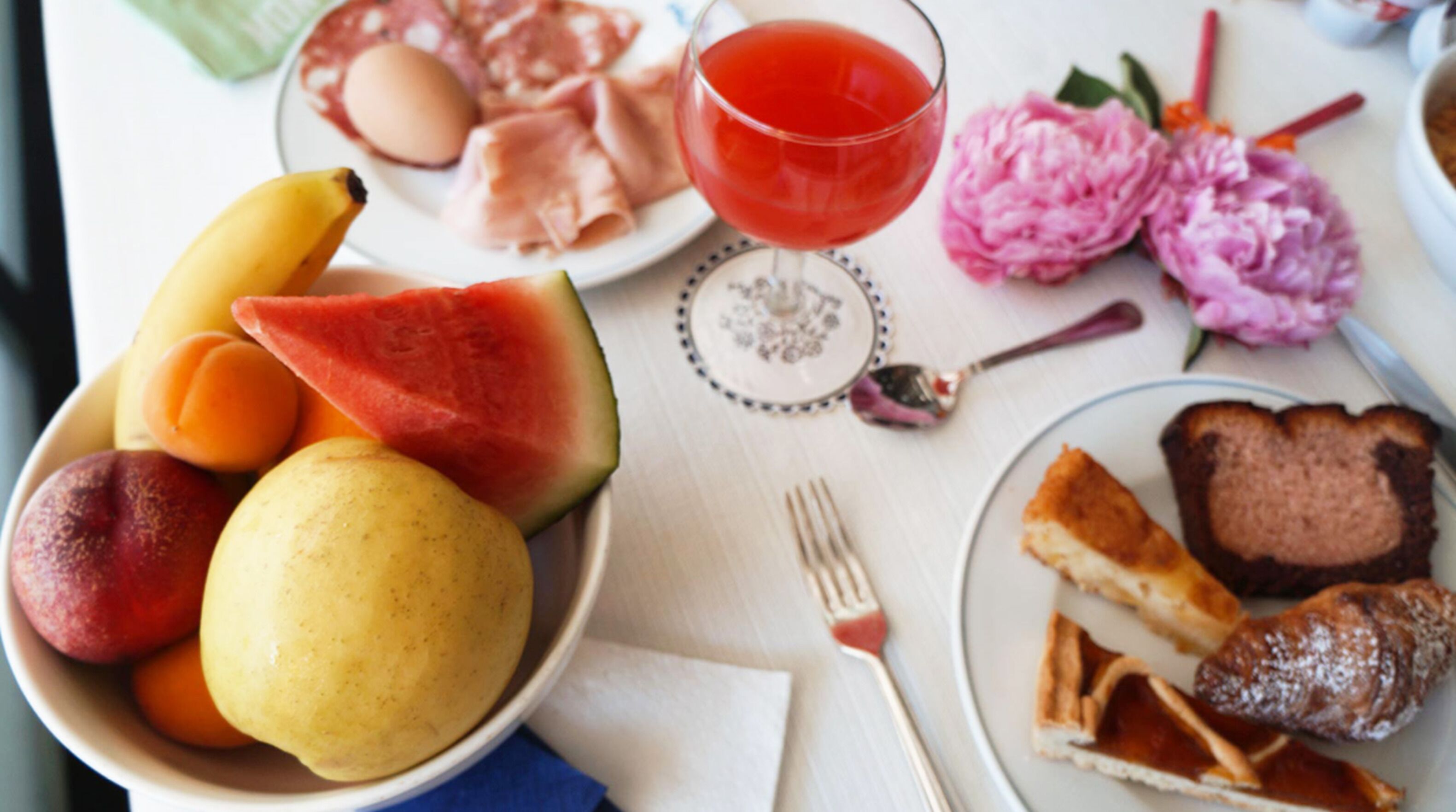 Una mesa con una fuente de pan en rebanadas, un cuenco de fruta, un vaso de bebida roja y una fuente de carne cortada.