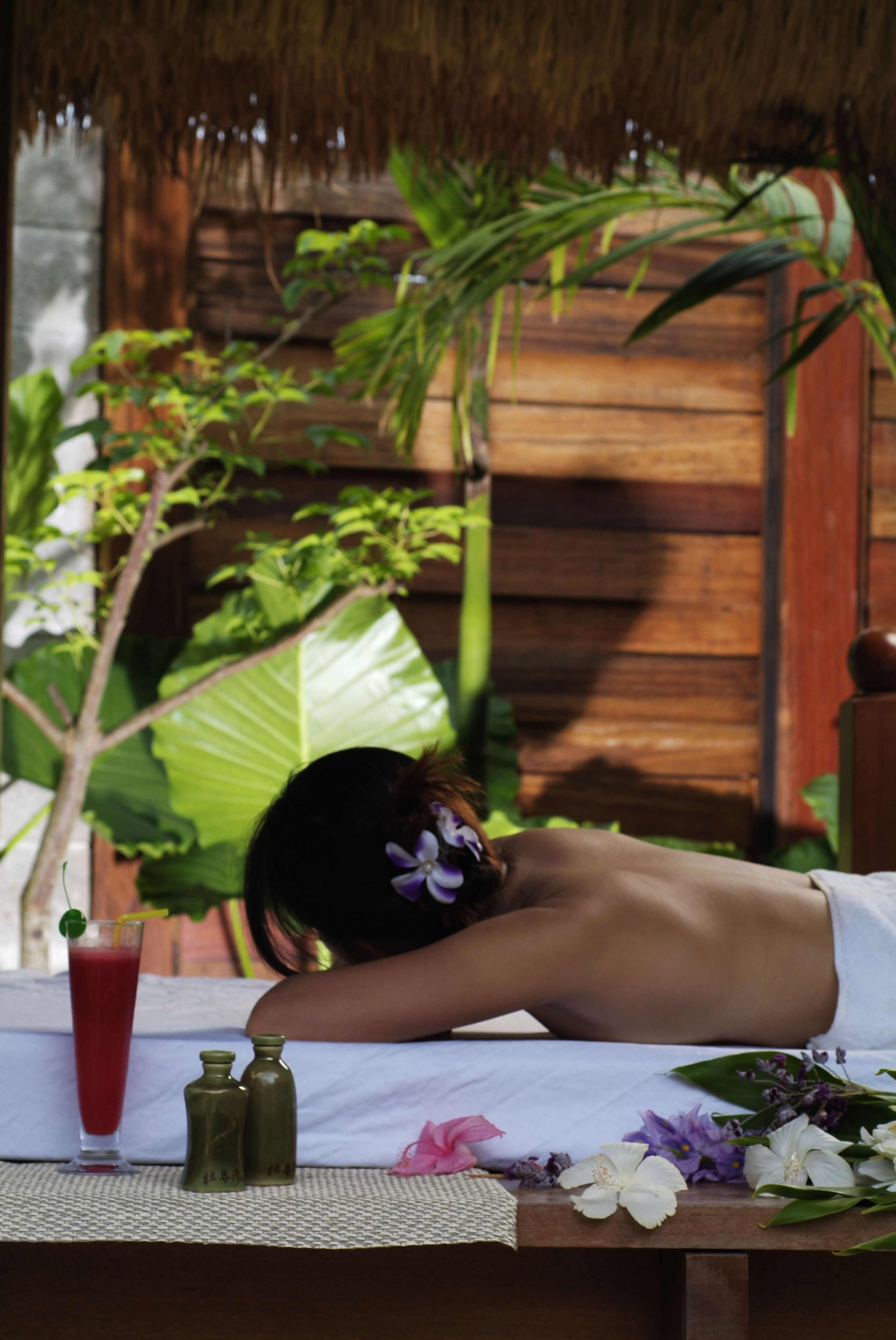 A person lying on a bed with a drink and flowers on a table.