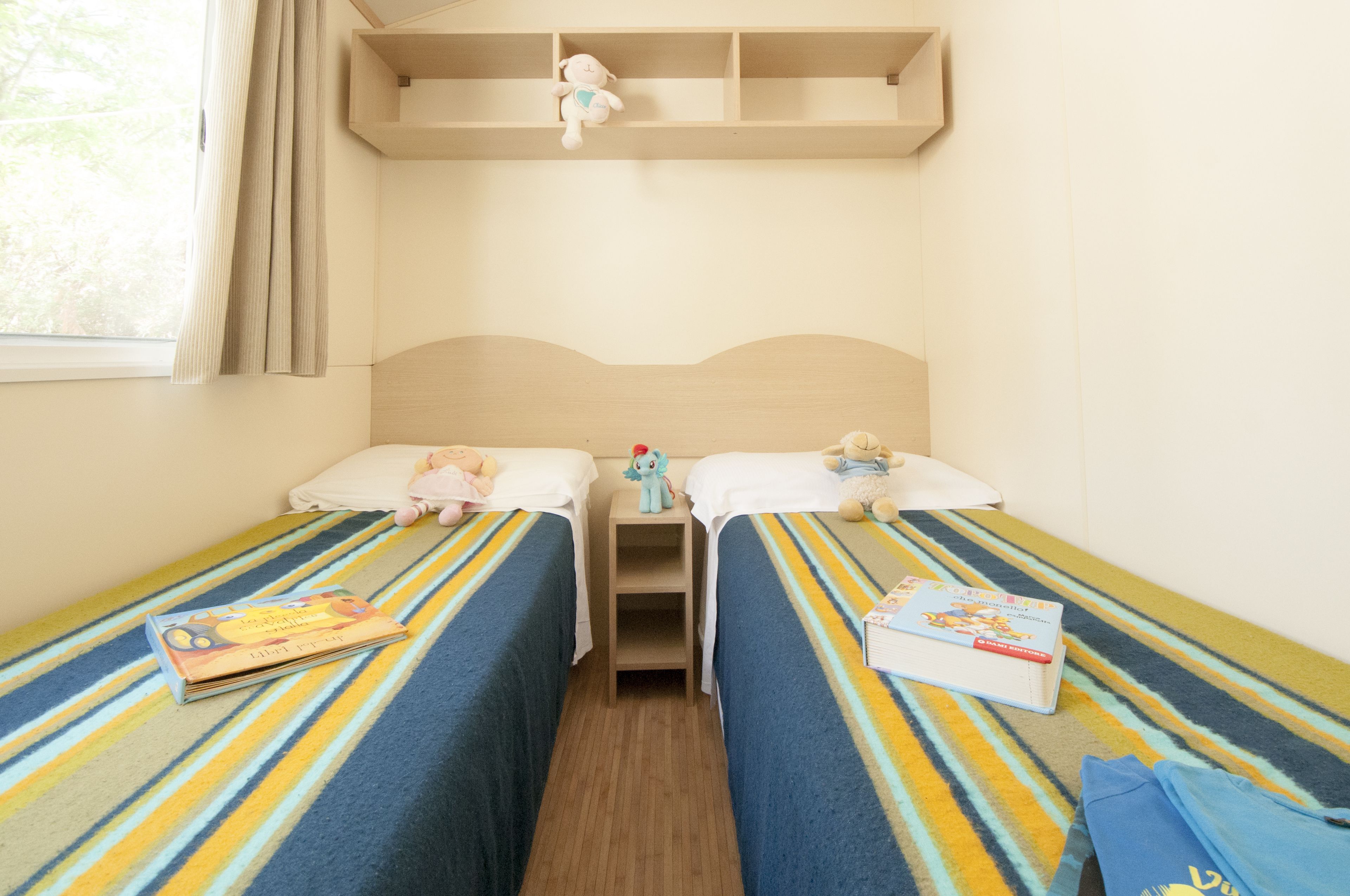 Ein Zimmer mit zwei Betten, jeweils mit gestreifter Bettdecke, einem kleinen Regal darüber und einem Fenster mit Vorhängen.