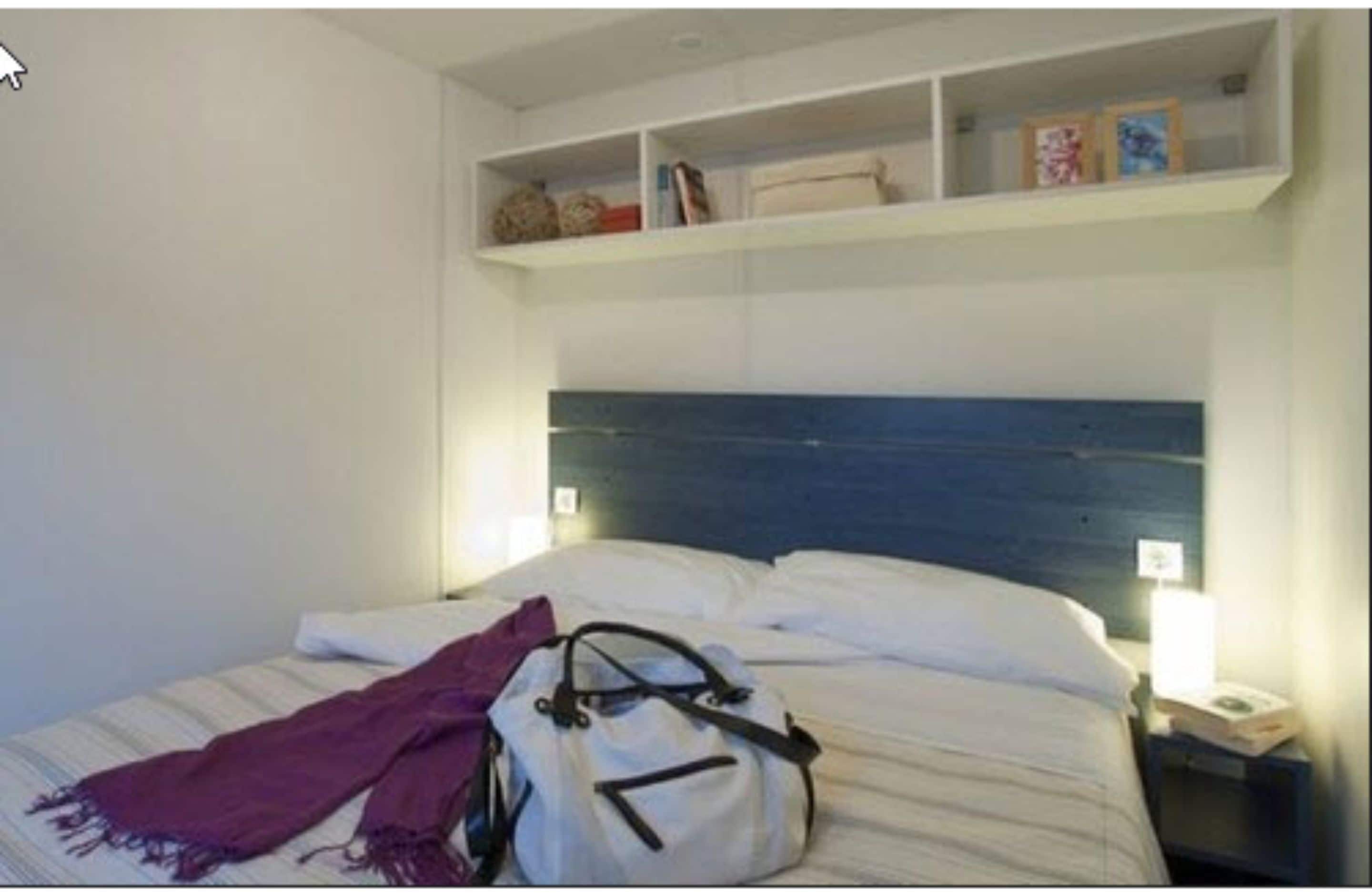 A hotel room with a bed, a nightstand, a shelf above the bed, and a bag on the bed.