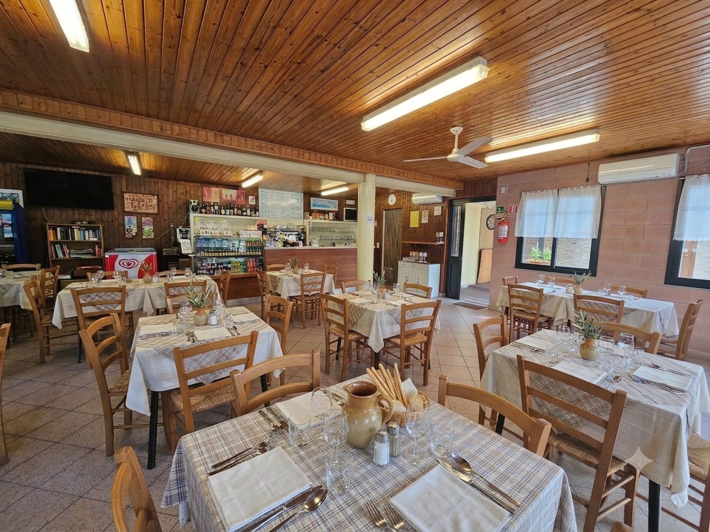A restaurant with wooden ceiling, tables set with white tablecloths, and a bar area with shelves of bottles.