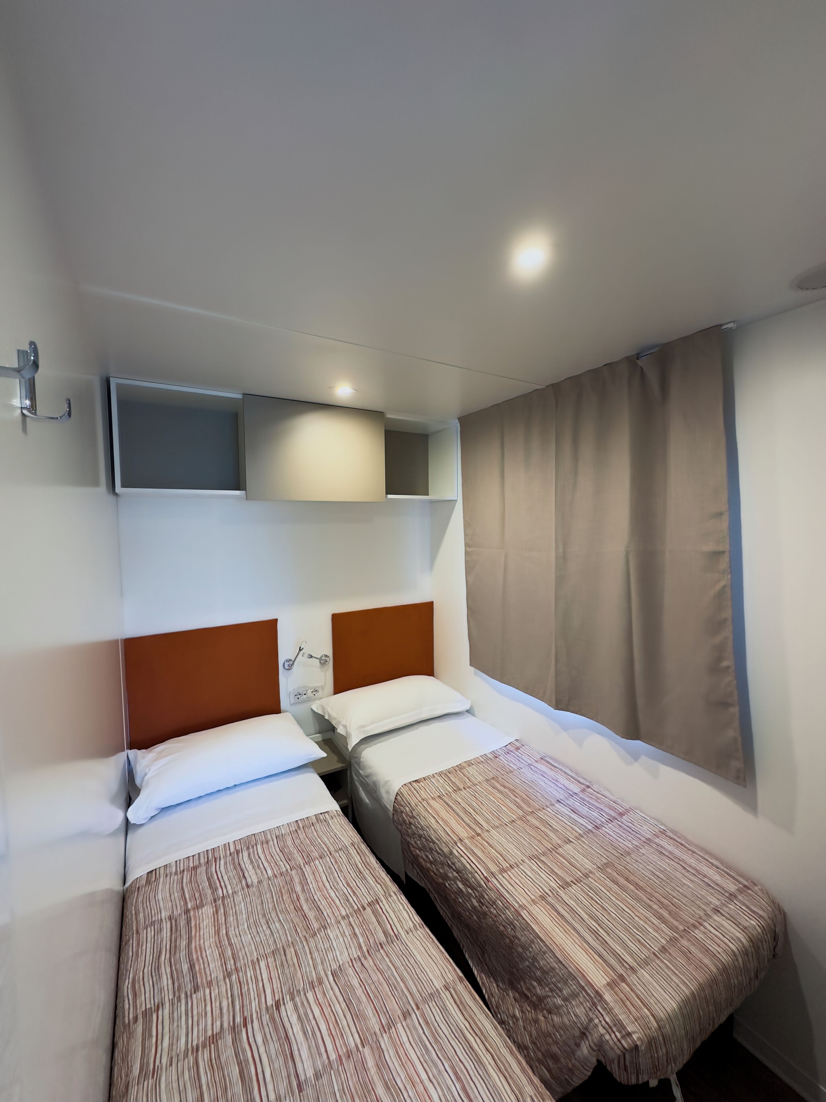 A hotel room with two single beds, a headboard, a shelf above the beds, a window with curtains, and a wall-mounted light.