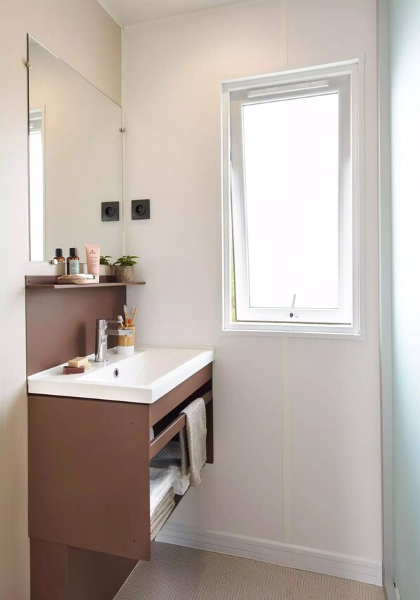 Un baño con lavamanos blanco, espejo y ventana. Hay un perchero y una repisa con artículos de tocador.