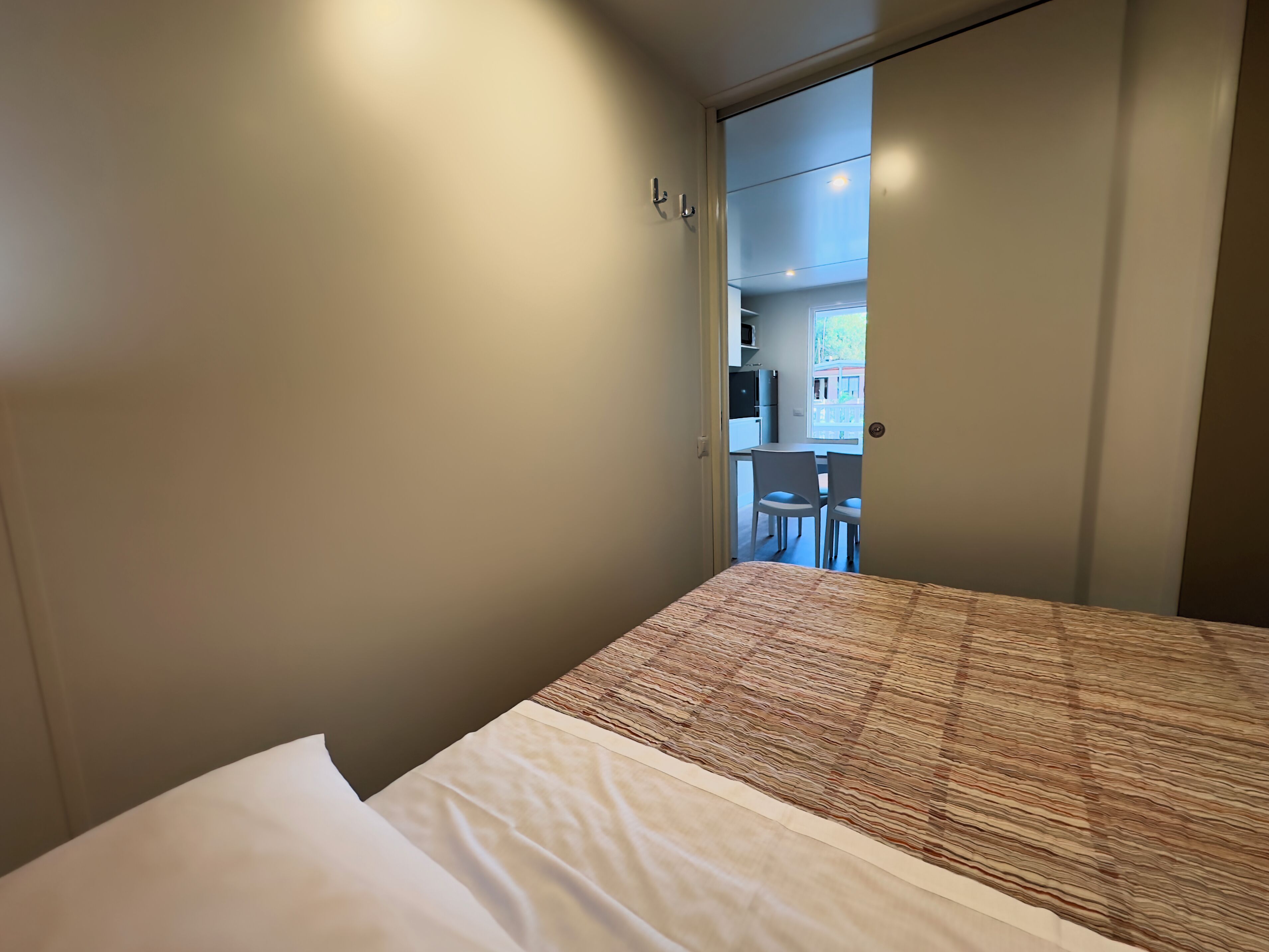 A bedroom with a bed, a door leading to a kitchenette, and a view of the outside.