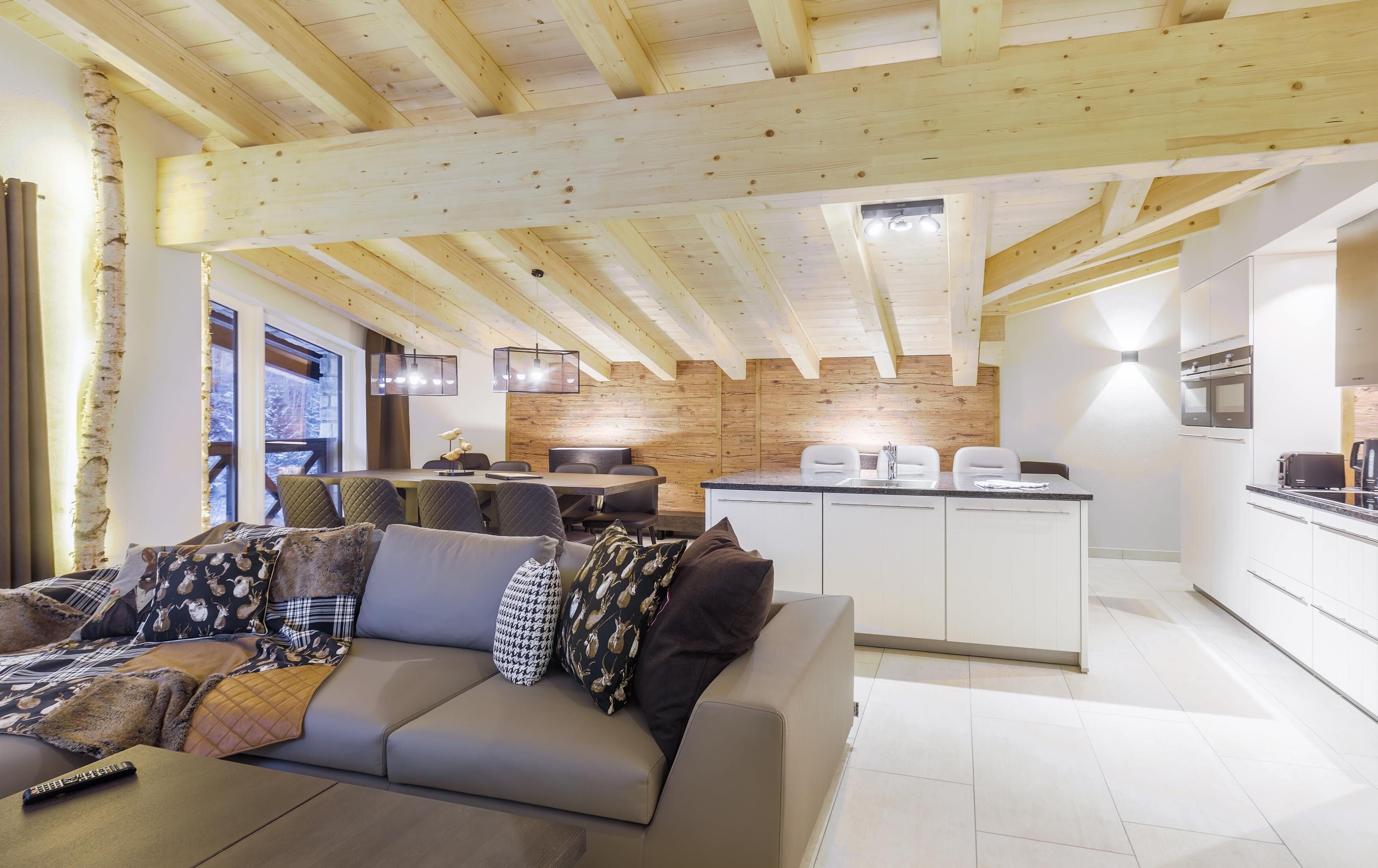 Une cuisine moderne avec des meubles blancs et un îlot central, un coin repas avec une table et des chaises en bois, et un espace salon confortable avec un canapé et des coussins.