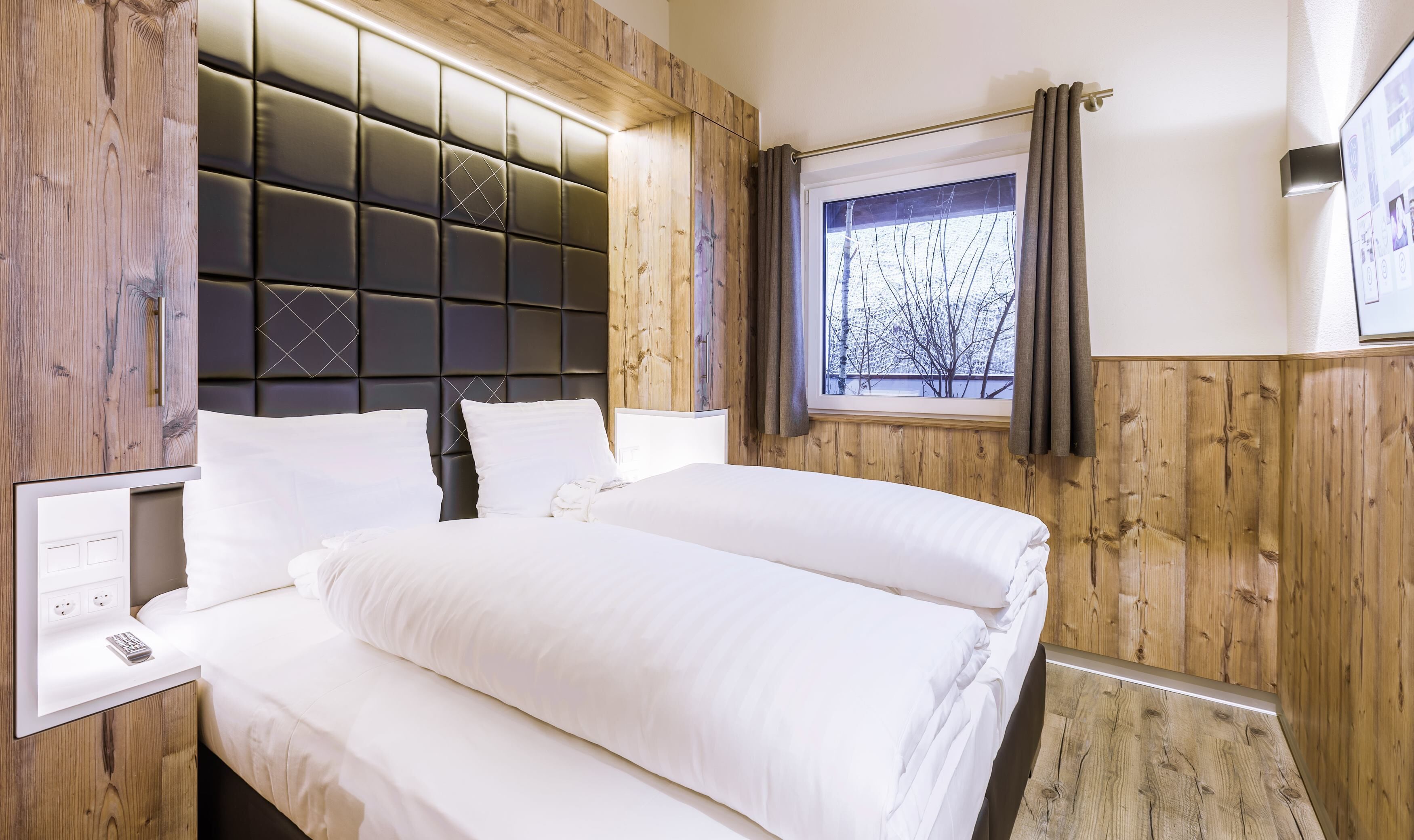 A hotel room with a wooden panel wall, a padded headboard, a large window with curtains, and a bedside table with a remote control.