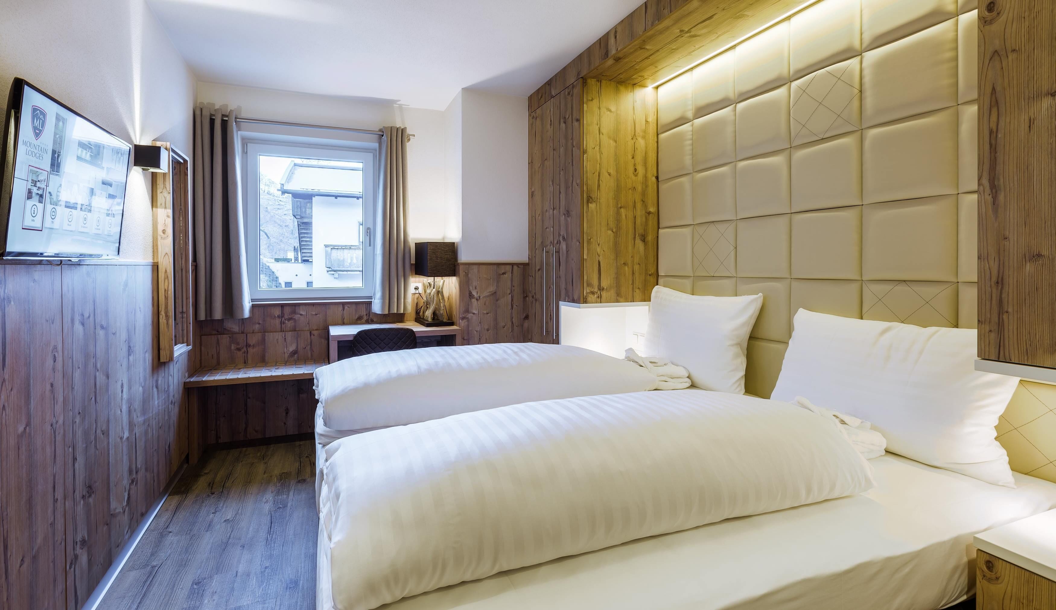 Habitación de hotel con dos camas, paneles de madera, un escritorio, una ventana con cortinas y un televisor montado.