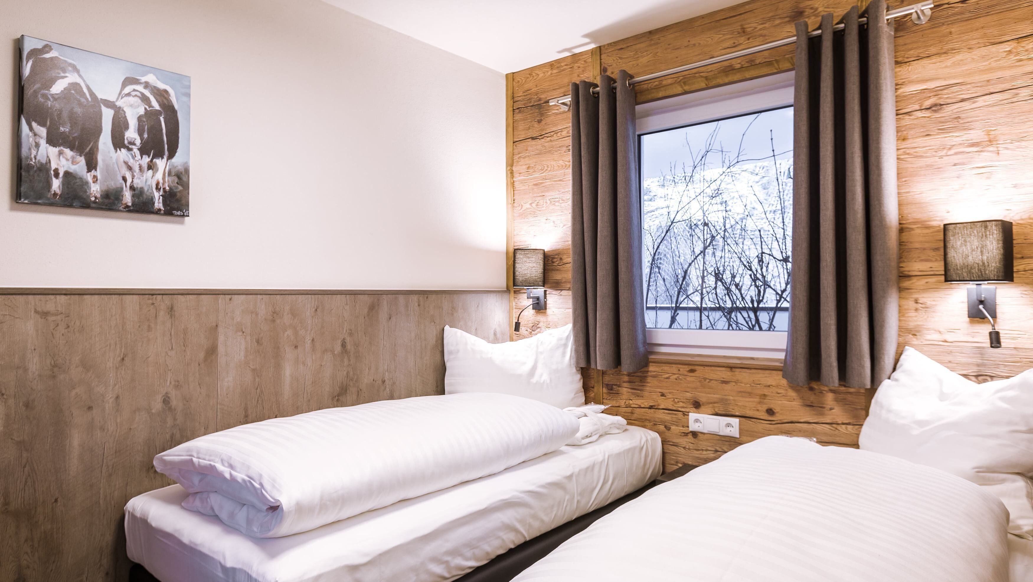 Habitación de hotel con dos camas, cabecero de madera, un cuadro de vacas, una ventana con cortinas y una lámpara de pared.