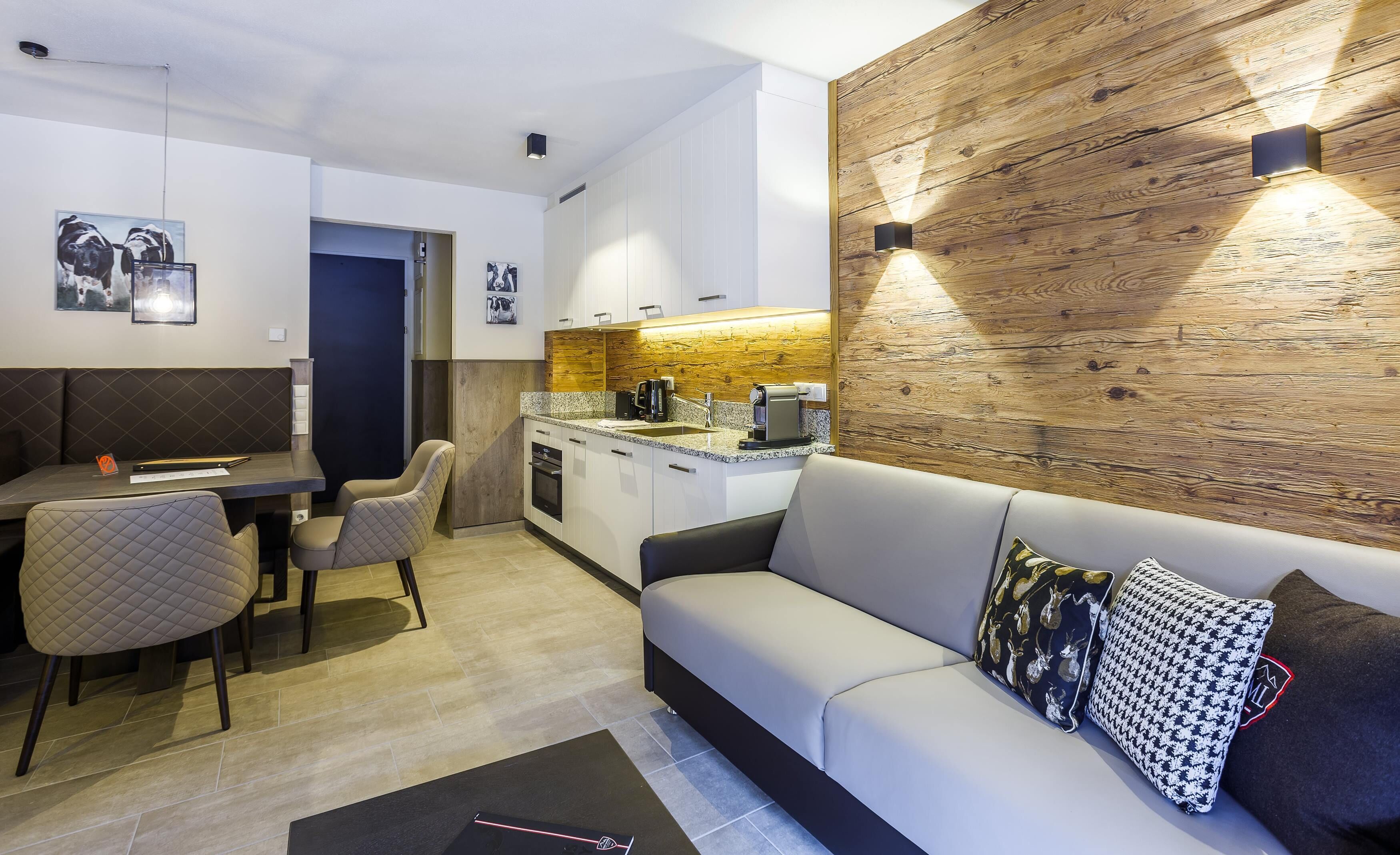 A modern living room with a sofa, a dining area with chairs, a kitchen with a countertop and appliances, and a wooden accent wall.