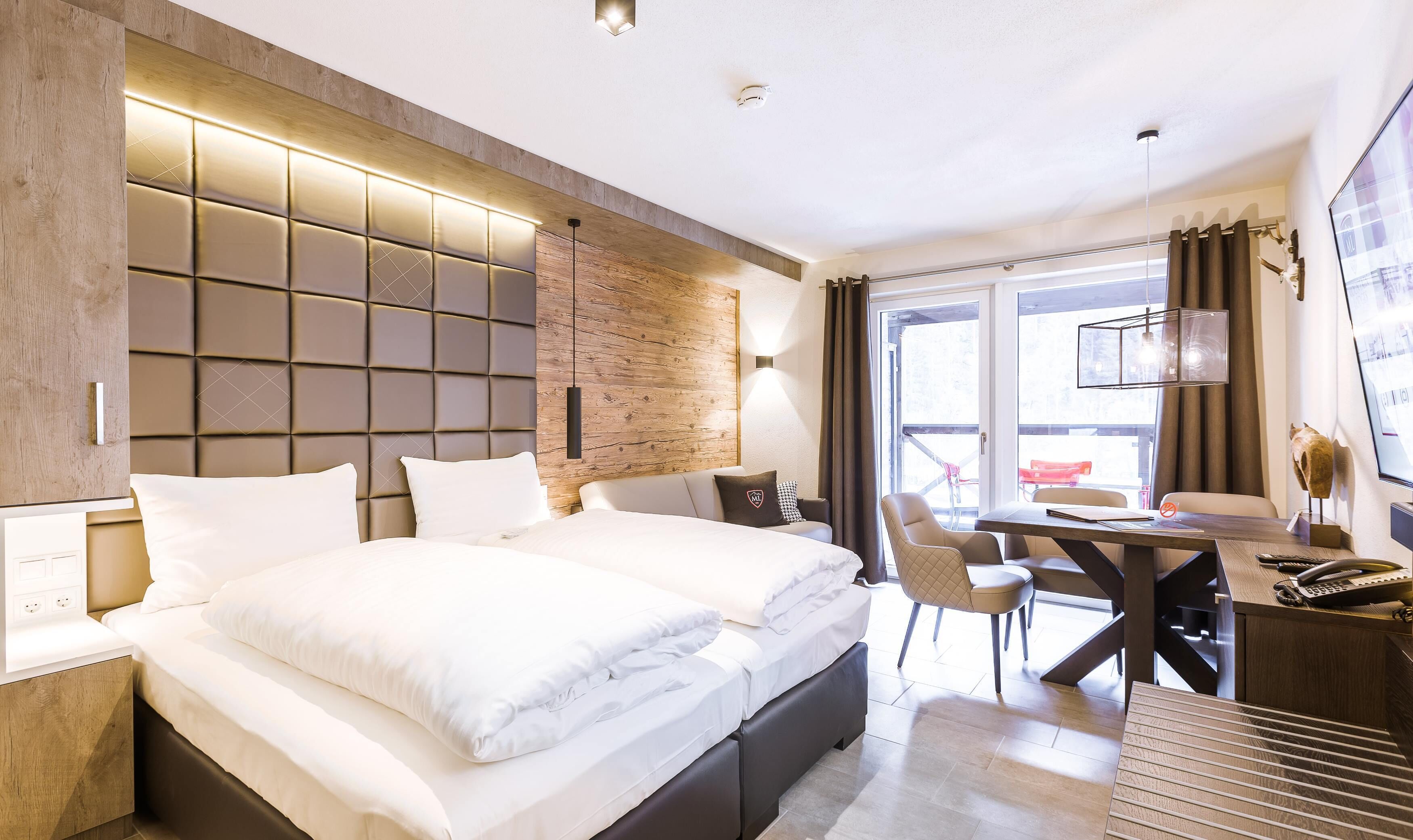 A modern hotel room with a large bed, a desk with a chair, and a view of the outdoors through a large window.