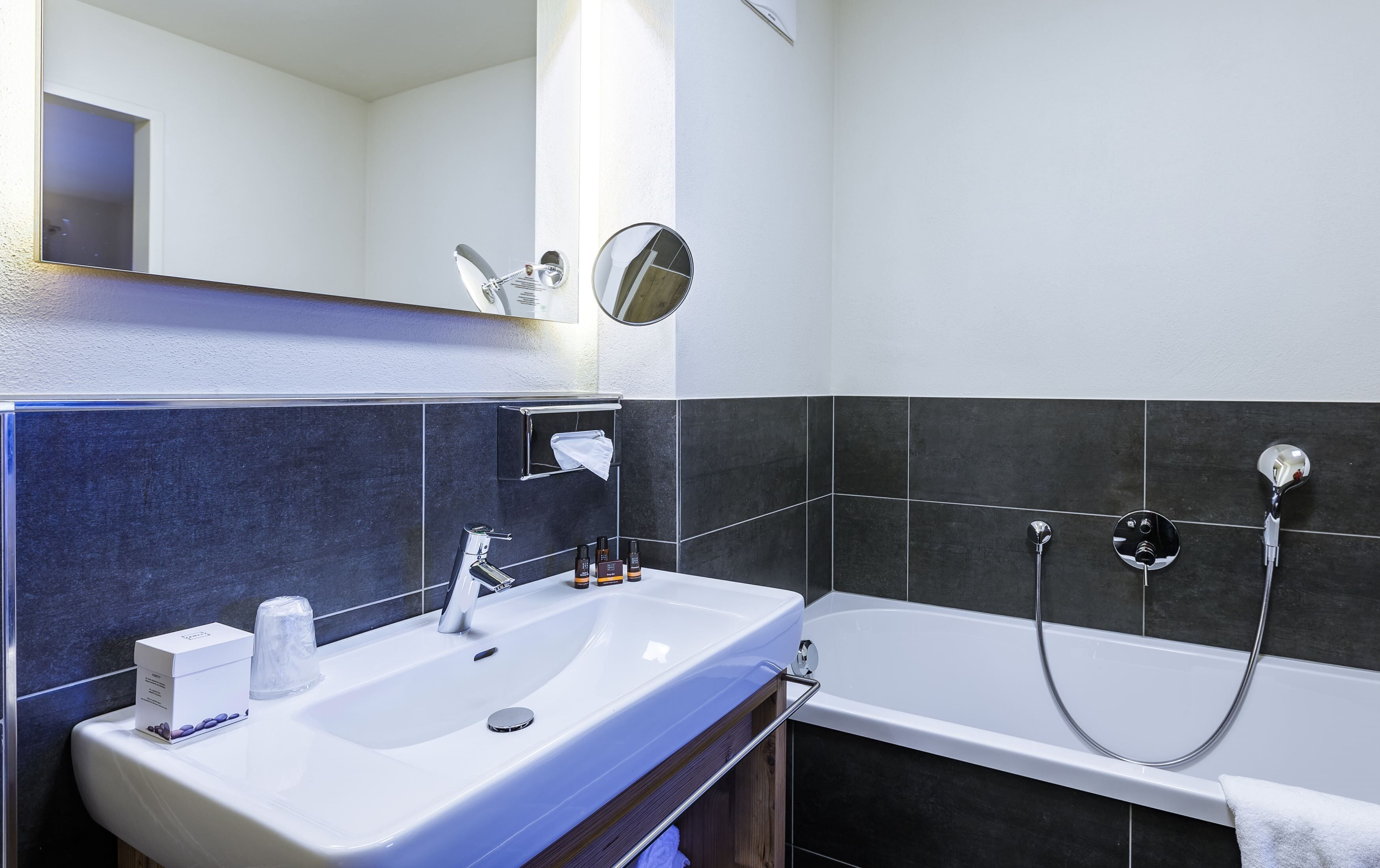 Ein modernes Badezimmer mit einem weißen Waschbecken, einem Spiegel, einer Handbrause und dunkel gefliesten Wänden.