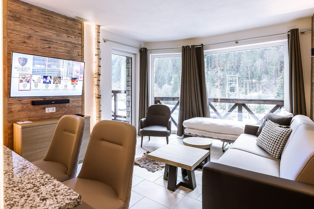 Un salon moderne avec un téléviseur fixé sur un mur en bois, un canapé, deux fauteuils, une table basse et une vue sur une forêt enneigée à travers de grandes fenêtres.