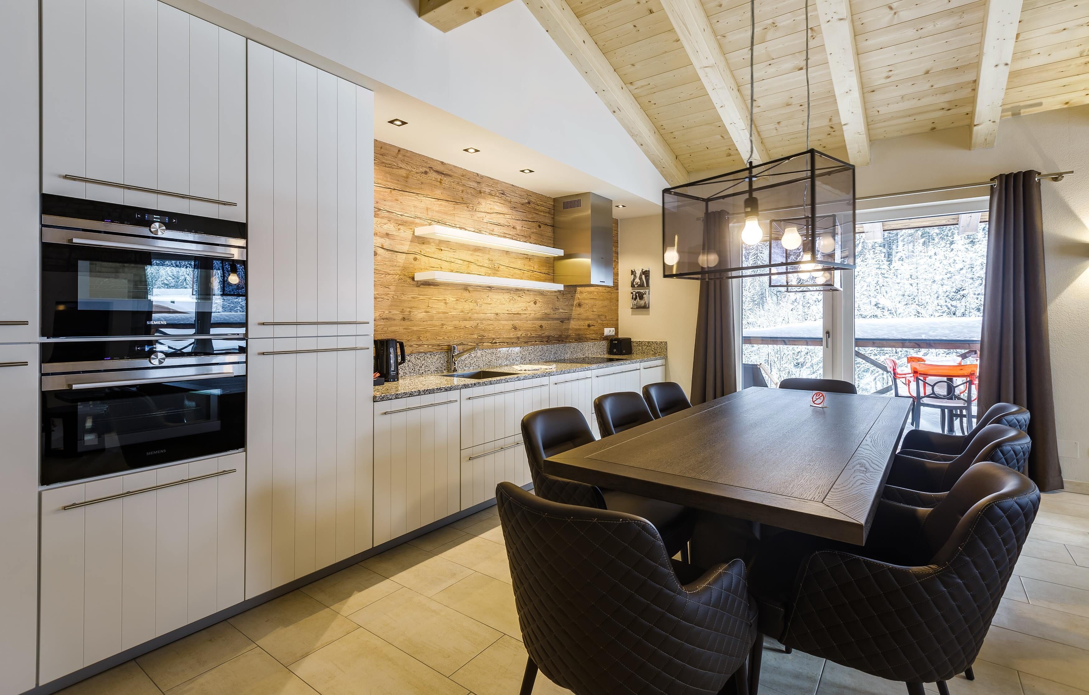 A modern kitchen with white cabinetry, a wooden accent wall, a built-in oven, and a dining area with a large table and chairs.