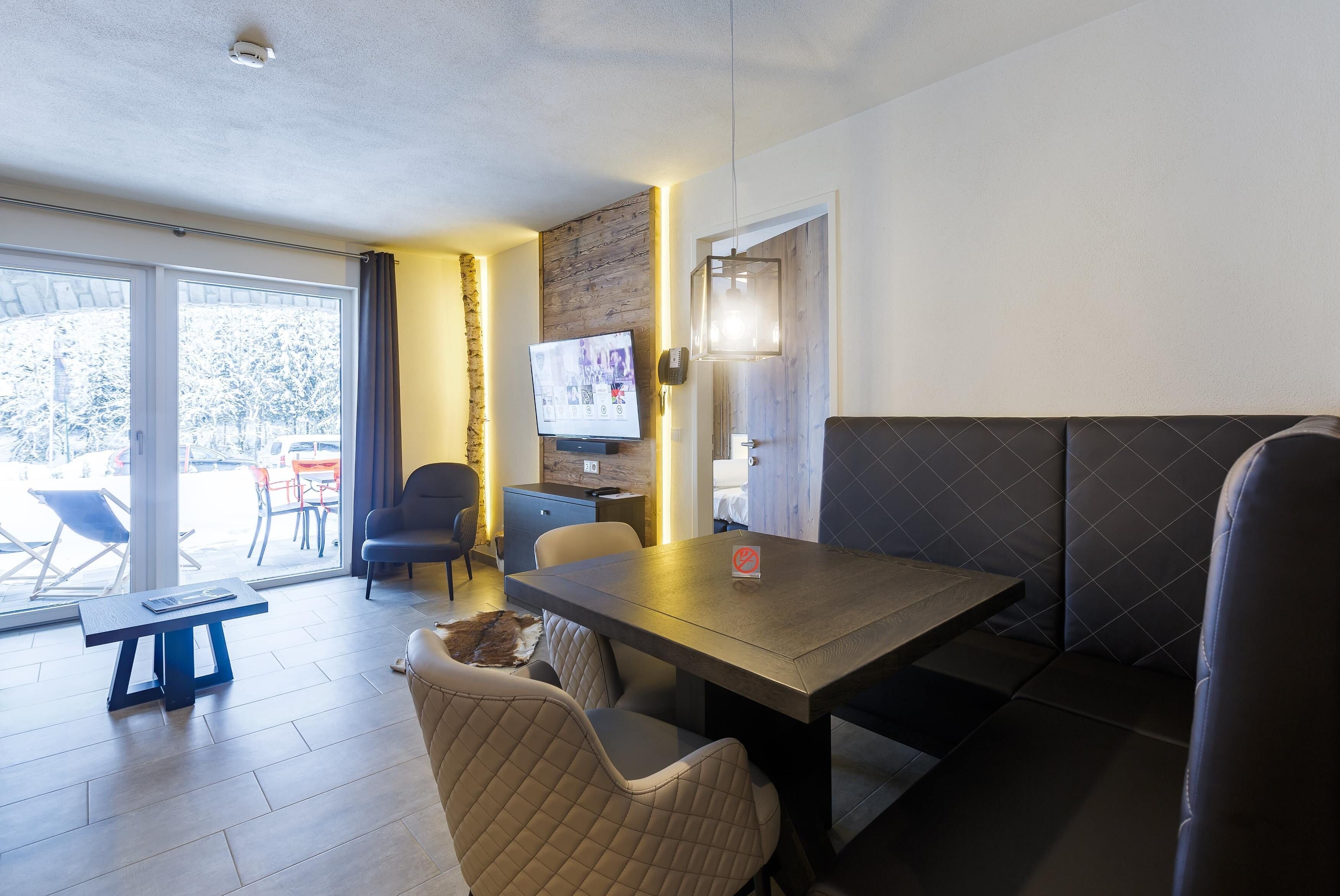 A modern room with a wooden table, cushioned seating, a television, and a large window with a view of outdoor seating.
