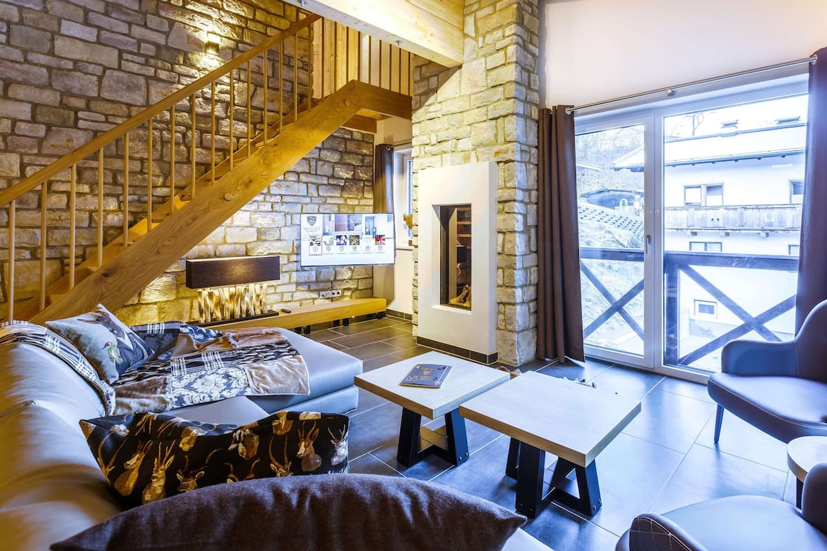Un salon chaleureux avec un mur en pierre, un escalier en bois, une cheminée et une grande fenêtre donnant sur un beau paysage.
