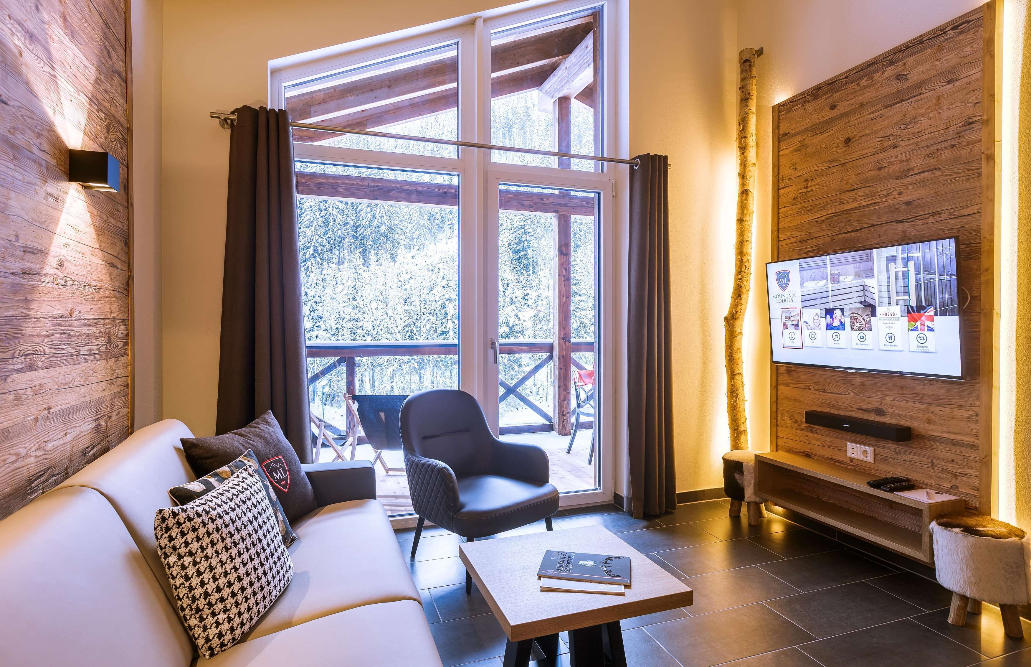 A modern living room with a sofa, armchair, and coffee table. A television is mounted on the wall, and a window provides a view of the outdoors.