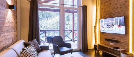 Una sala moderna con un sofá, un sillón y una mesa de centro. Un televisor está montado en la pared, y una ventana ofrece vistas al exterior.