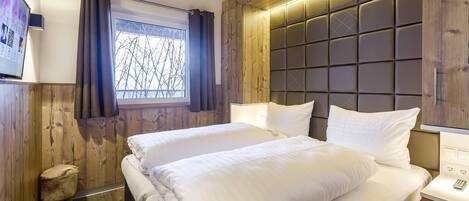 Quarto de hotel com uma cama grande, painéis de madeira, janela com cortinas e cabeceira moderna.