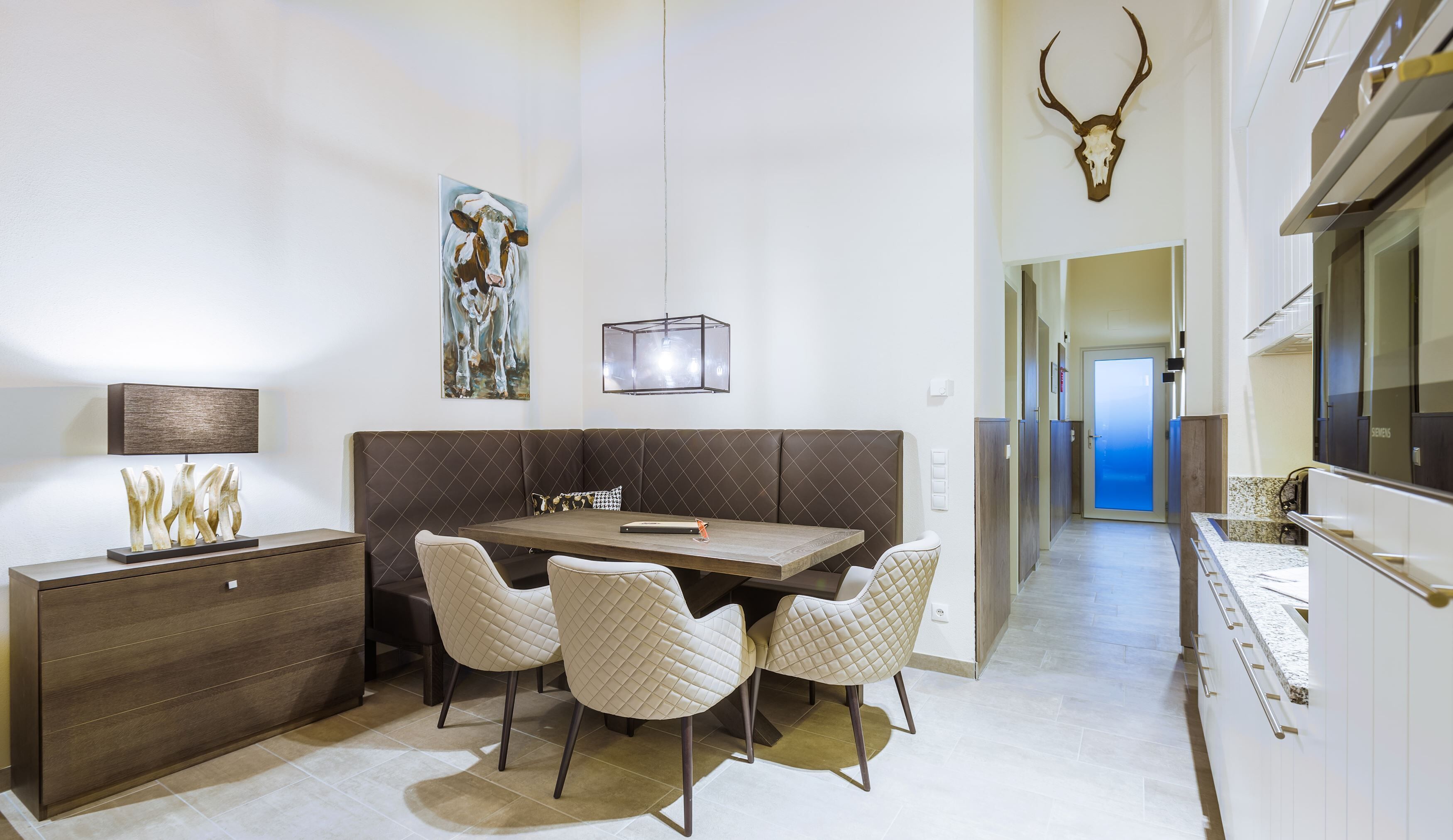 Un espace de salle à manger moderne avec une table en bois, des chaises rembourrées, un tableau et un crâne de cerf accroché au mur.