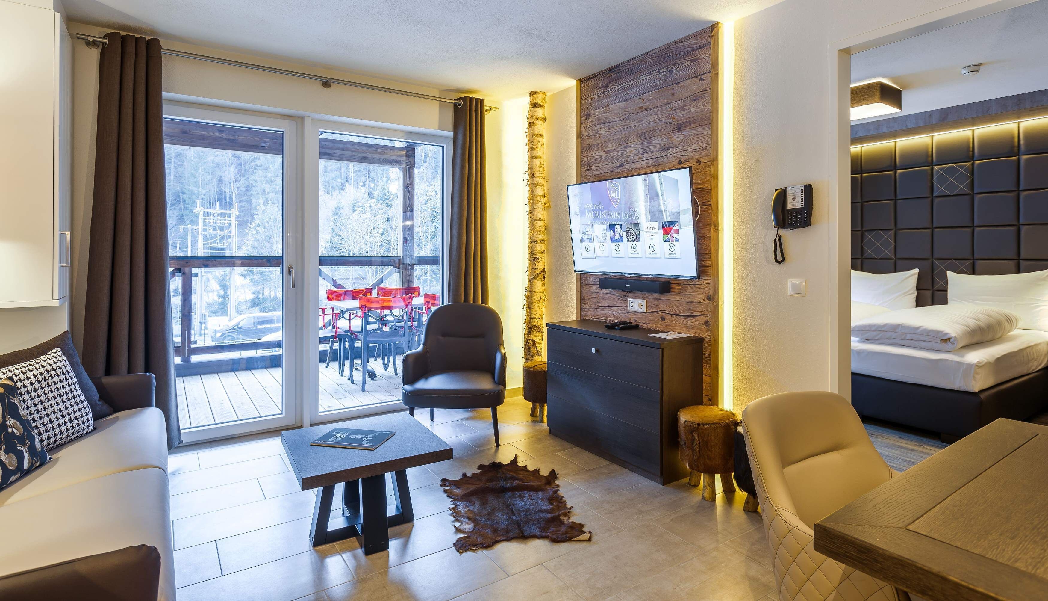 Een moderne hotelkamer met een grote schuifglazen deur naar een terras, een flatscreen-tv bevestigd aan een houten muur, een bed met witte lakens en een leren bank.