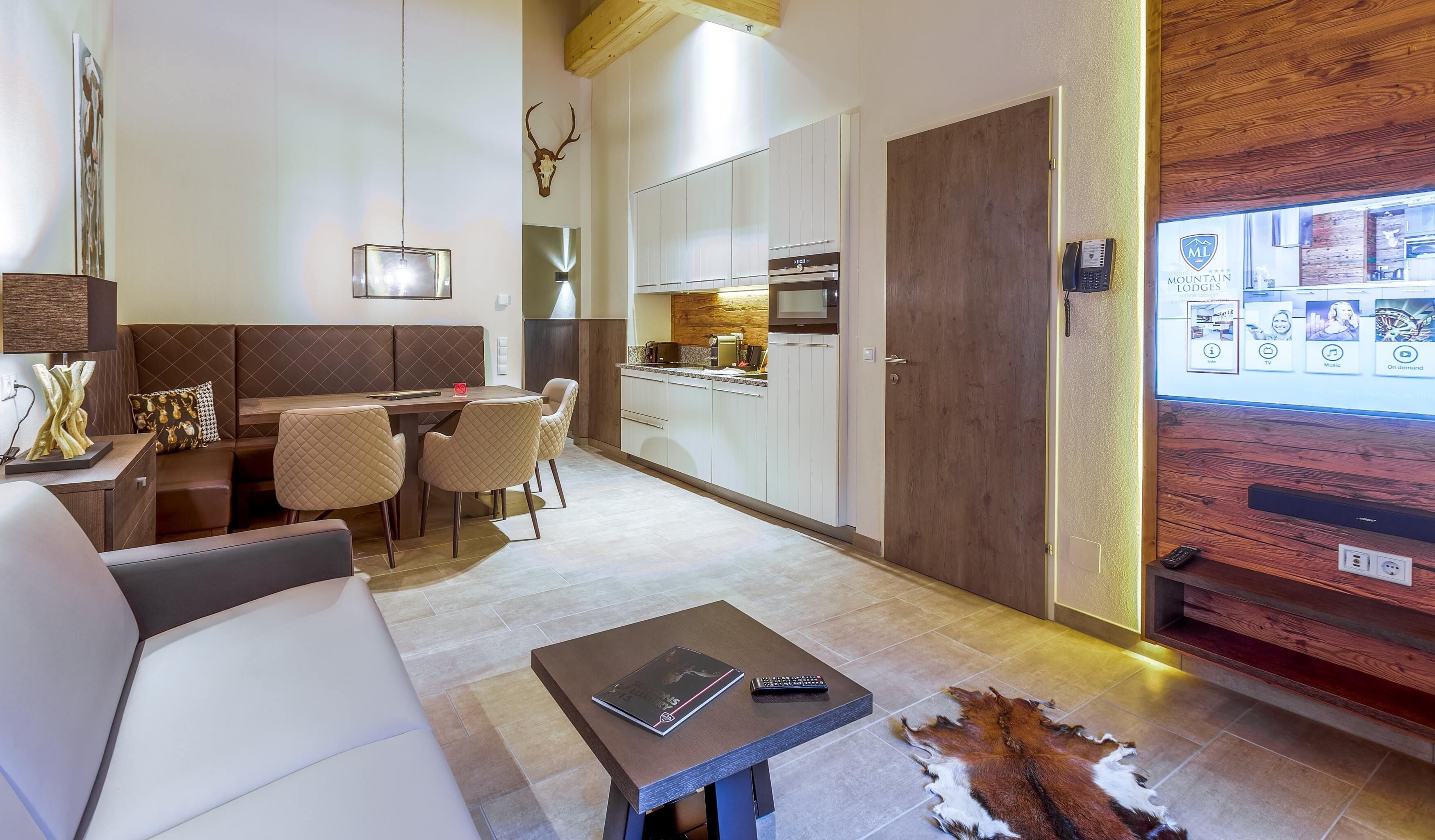 Una sala de estar moderna con un sofá, una mesa de comedor con sillas, una zona de cocina con armarios y electrodomésticos, y un televisor montado que muestra una interfaz de reserva de hotel.