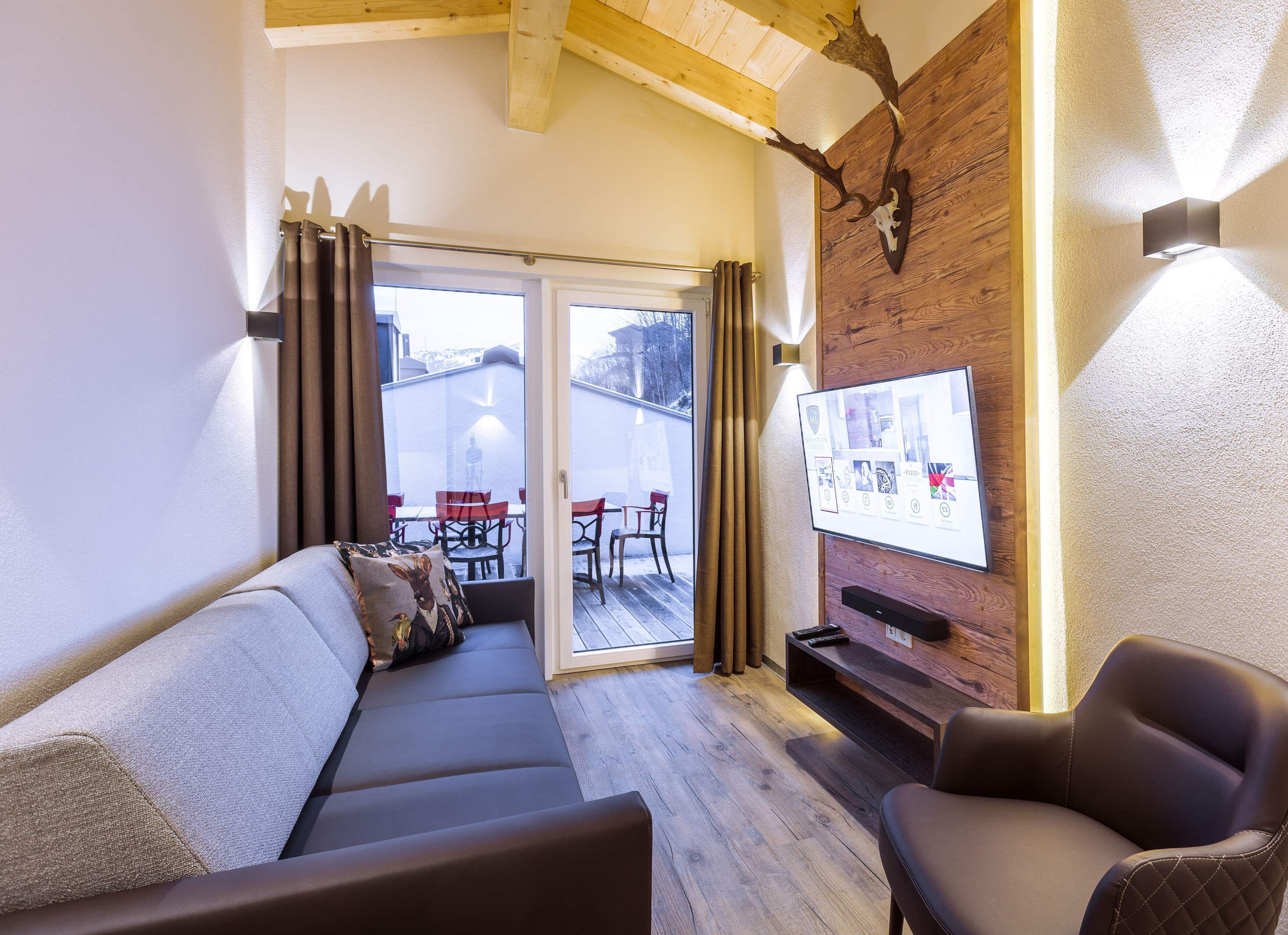 A cozy living room with a sofa, a chair, a TV mounted on a wooden wall, and a sliding glass door leading to an outdoor area with chairs.