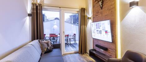 Un salon chaleureux avec un canapé, un fauteuil, un téléviseur fixé à un mur en bois et une porte-fenêtre coulissante donnant sur un espace extérieur aménagé avec des chaises.