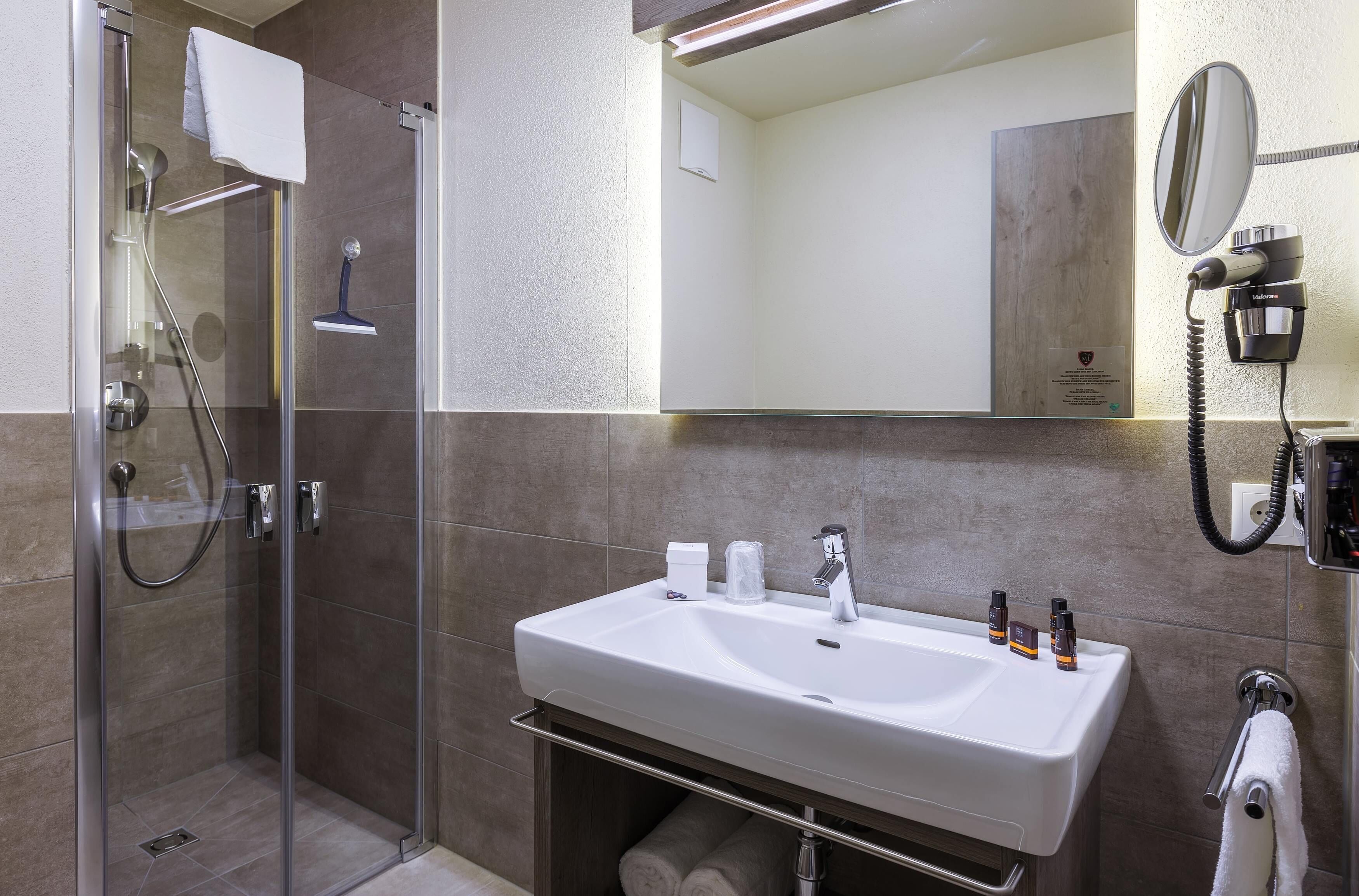 Un baño moderno con ducha de vidrio, un lavamanos blanco con espejo y artículos de aseo sobre el lavamanos.