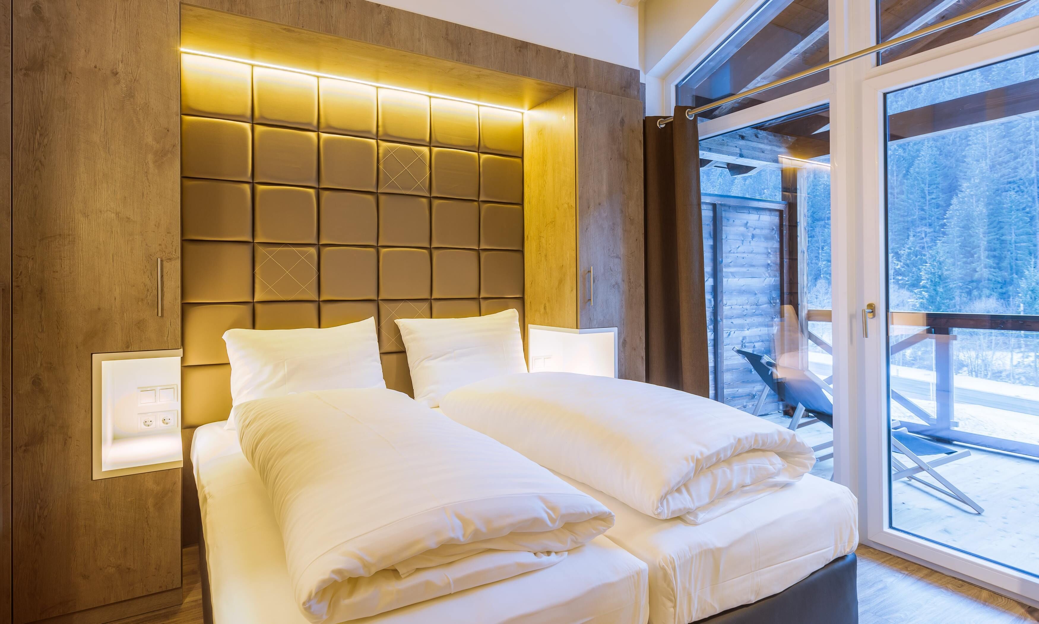 A modern bedroom with a wooden headboard, a large window with a view of a snowy landscape, and a neatly made bed with white linens.