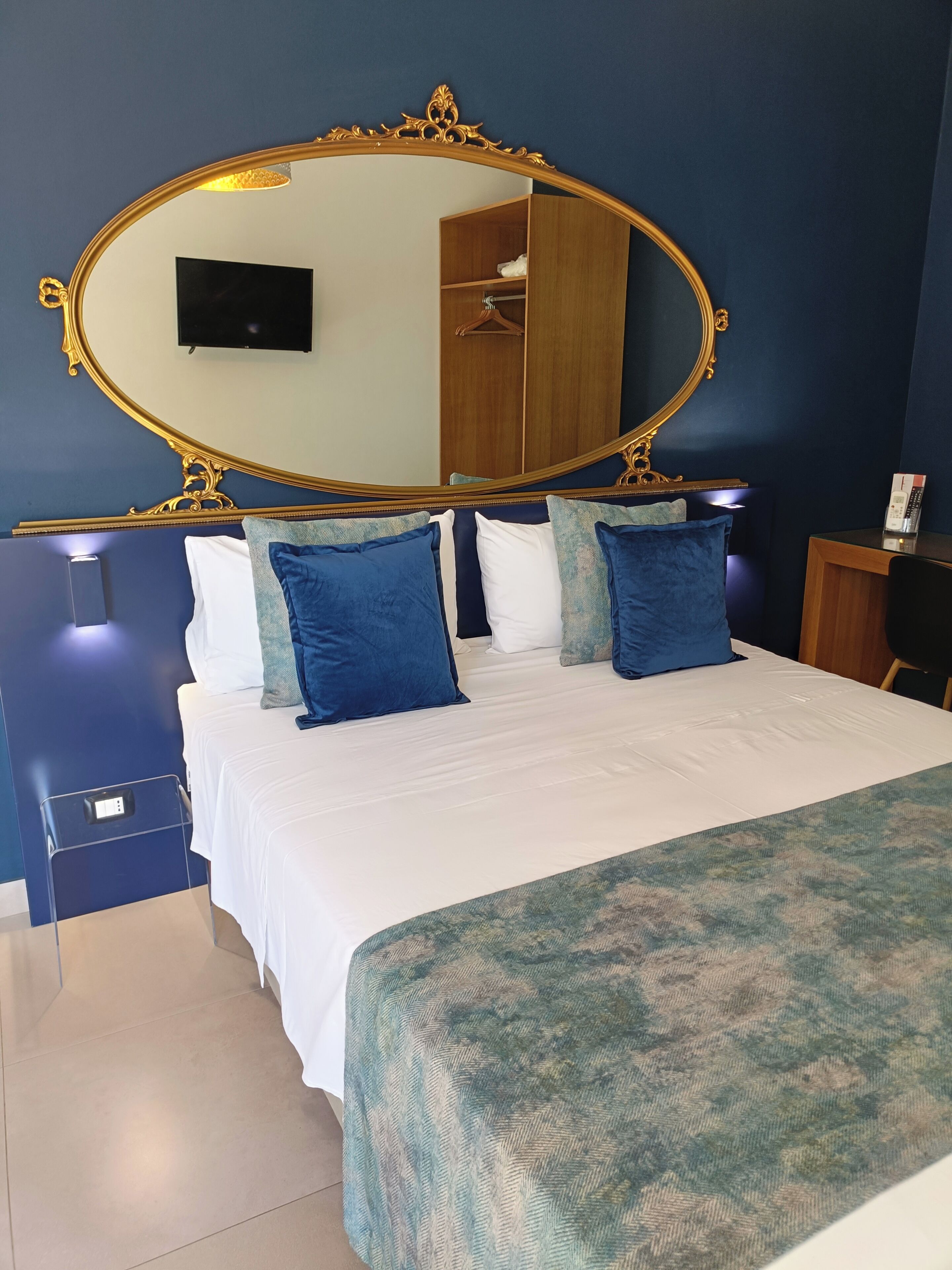 Une chambre à coucher avec un grand miroir décoratif, un lit avec une literie bleue et blanche, une table de chevet avec un téléphone et une armoire en arrière-plan.