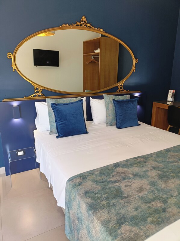 A bedroom with a large ornate mirror, a bed with blue and white bedding, a bedside table with a phone, and a wardrobe in the background.