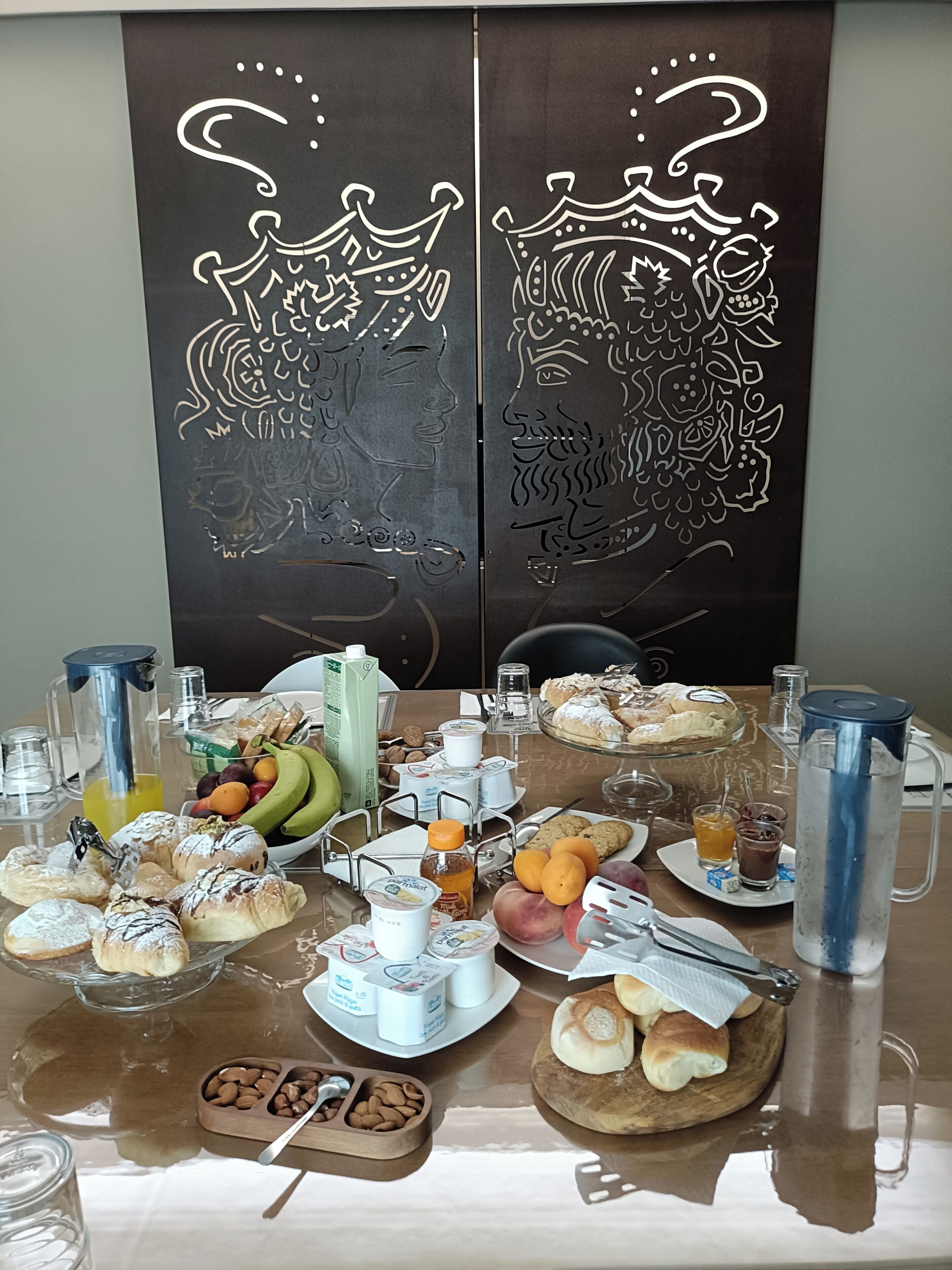 A breakfast buffet with various food items and beverages, a coffee maker, and a decorative panel with a crown design.
