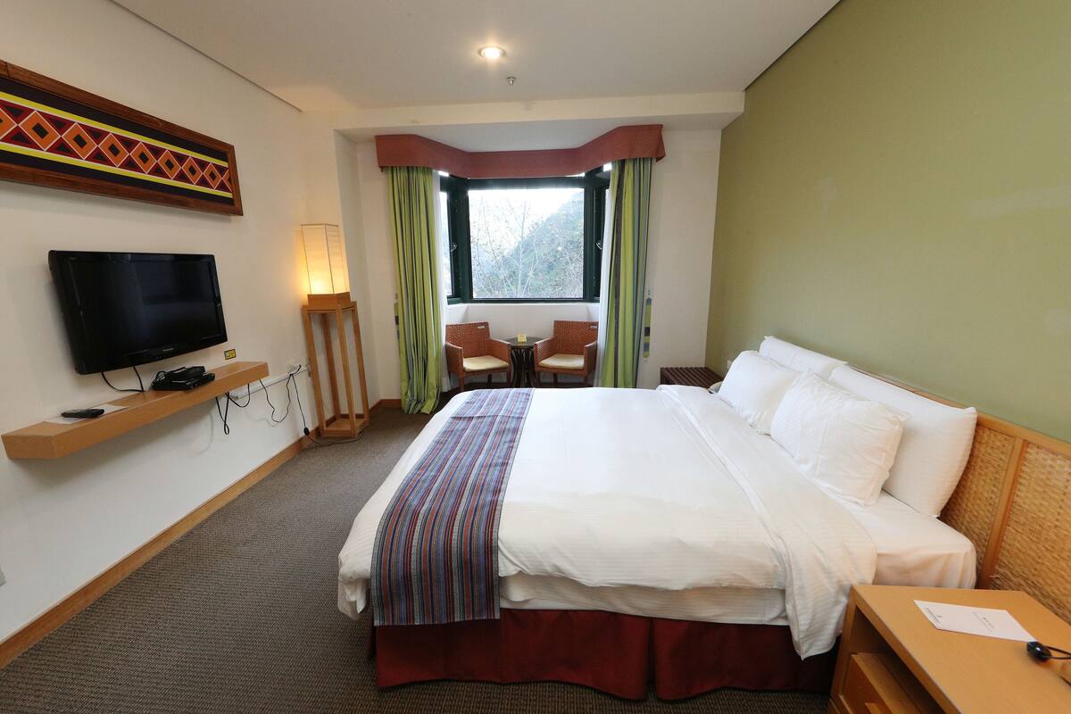 A hotel room with a large bed, a television on a shelf, a window with curtains, and a small table with chairs.