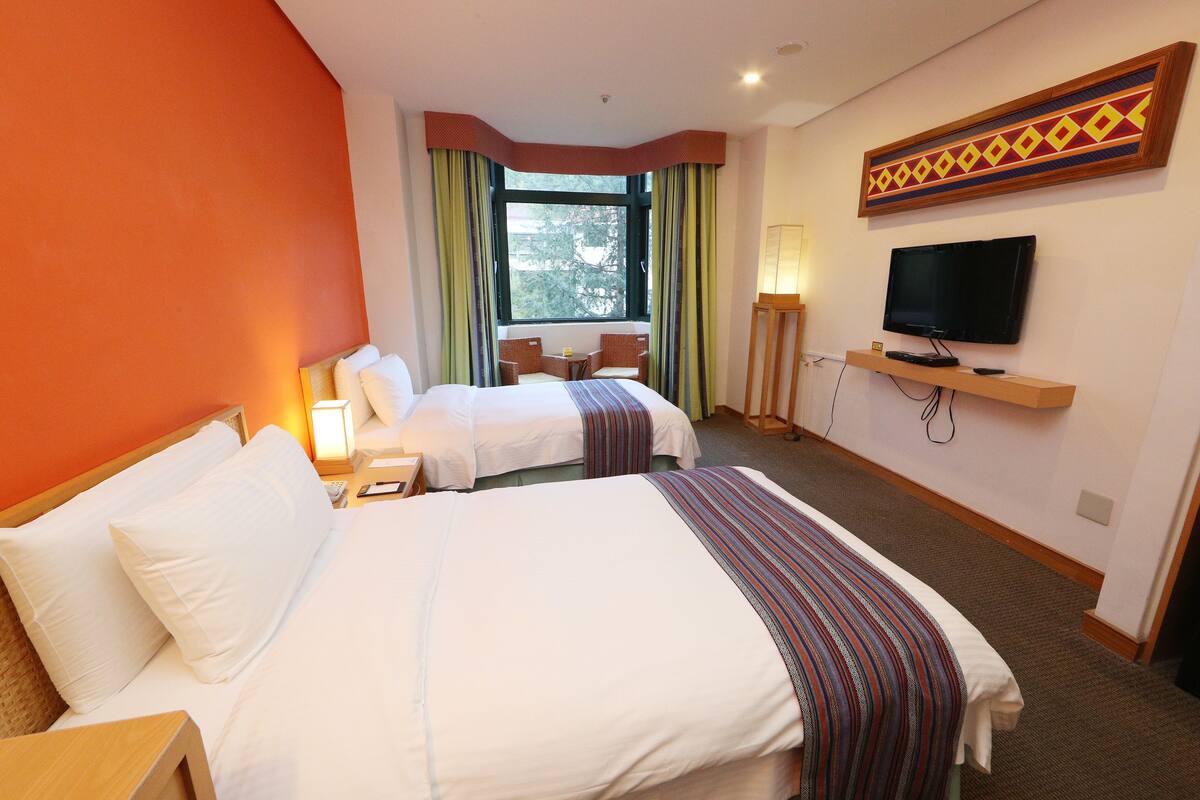 A hotel room with two beds, a TV, a desk, and a window with curtains.