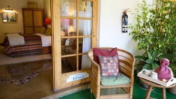 Una habitación con una cama, una silla de mimbre, una mesita con una bandeja y una planta en maceta.