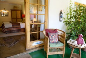 Una habitación con una cama, una silla de mimbre, una mesita con una bandeja y una planta en maceta.