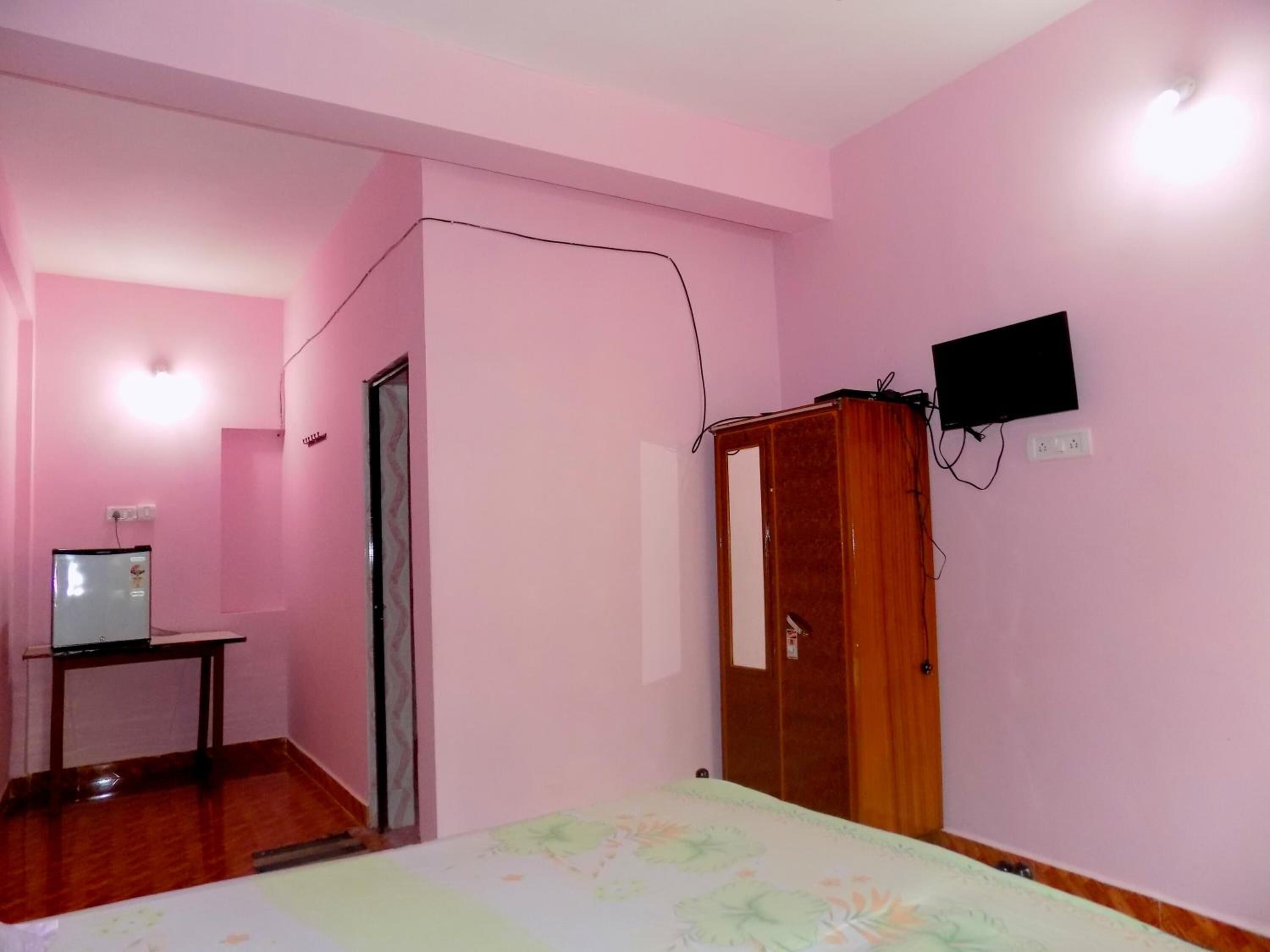 Una stanza dalle pareti rosa con un letto, un'armadio in legno, un tavolino con un televisore e una porta.