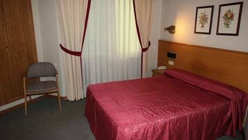 A hotel room with a bed, a chair, a wooden wardrobe, and a nightstand with a lamp.