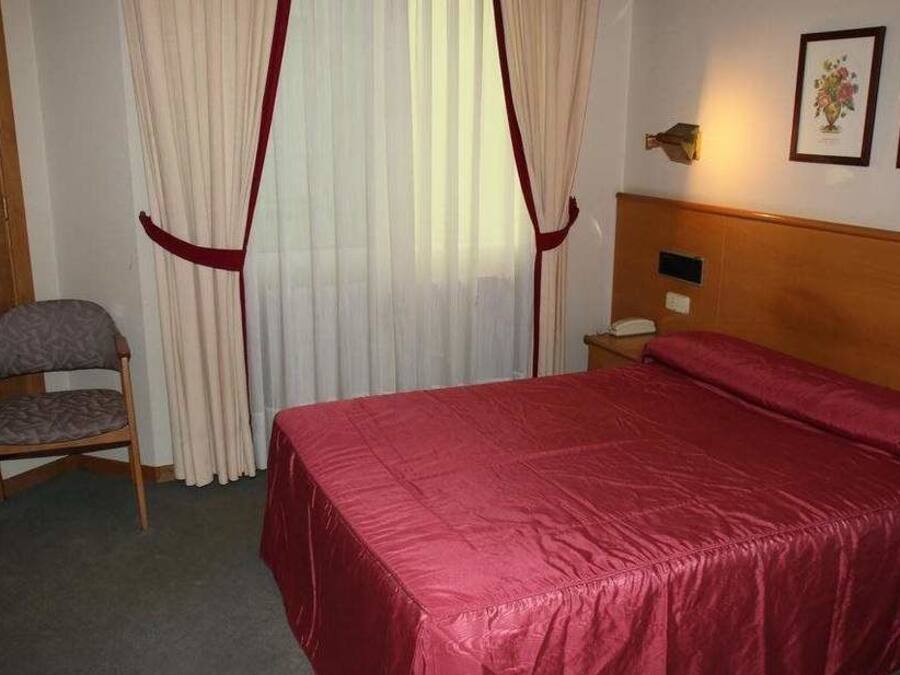 A hotel room with a bed, a chair, a wooden wardrobe, and a nightstand with a lamp.